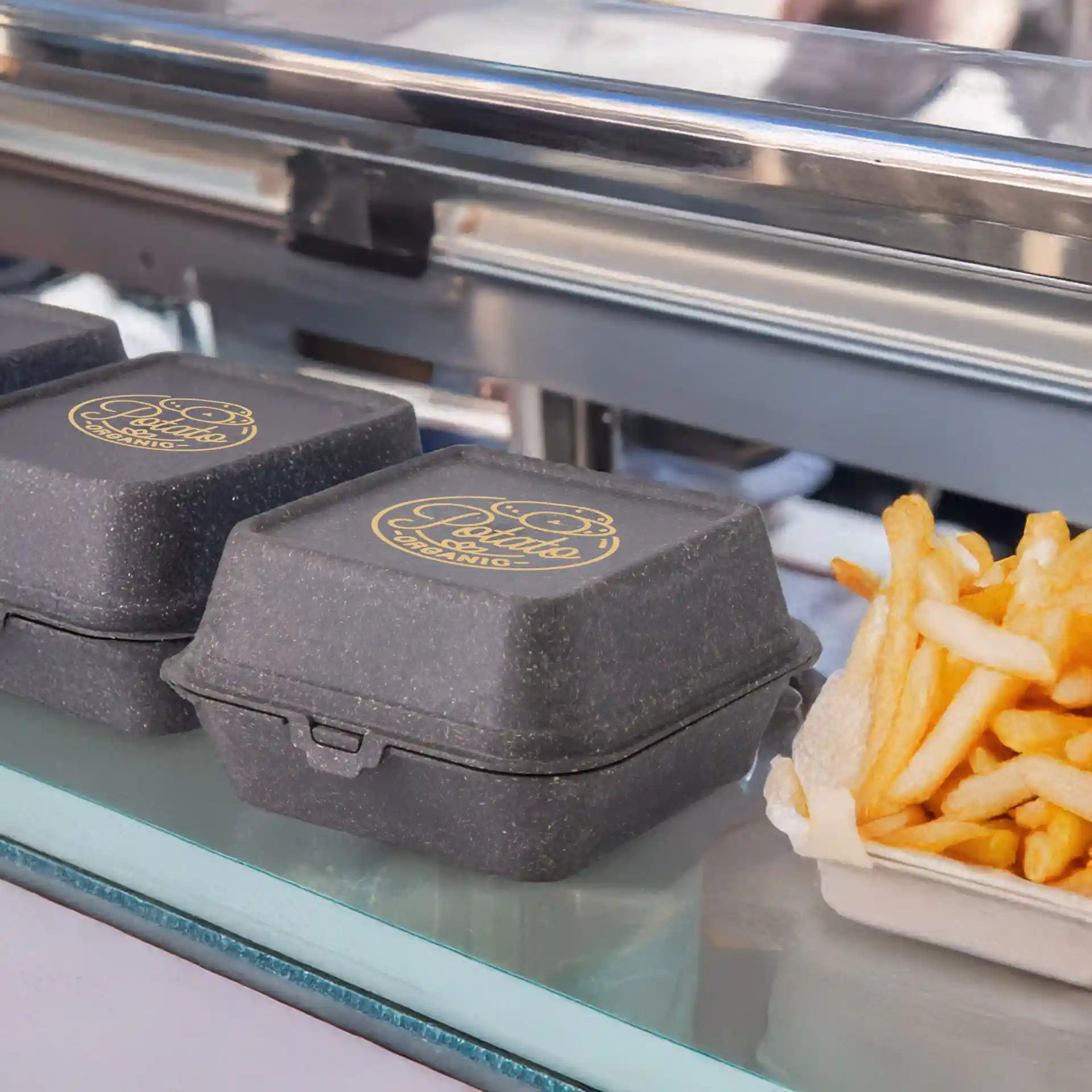 Trois boîtes à burger réutilisables grises imprimées avec un logo sur le dessus, placées à côté d'une portion de frites.