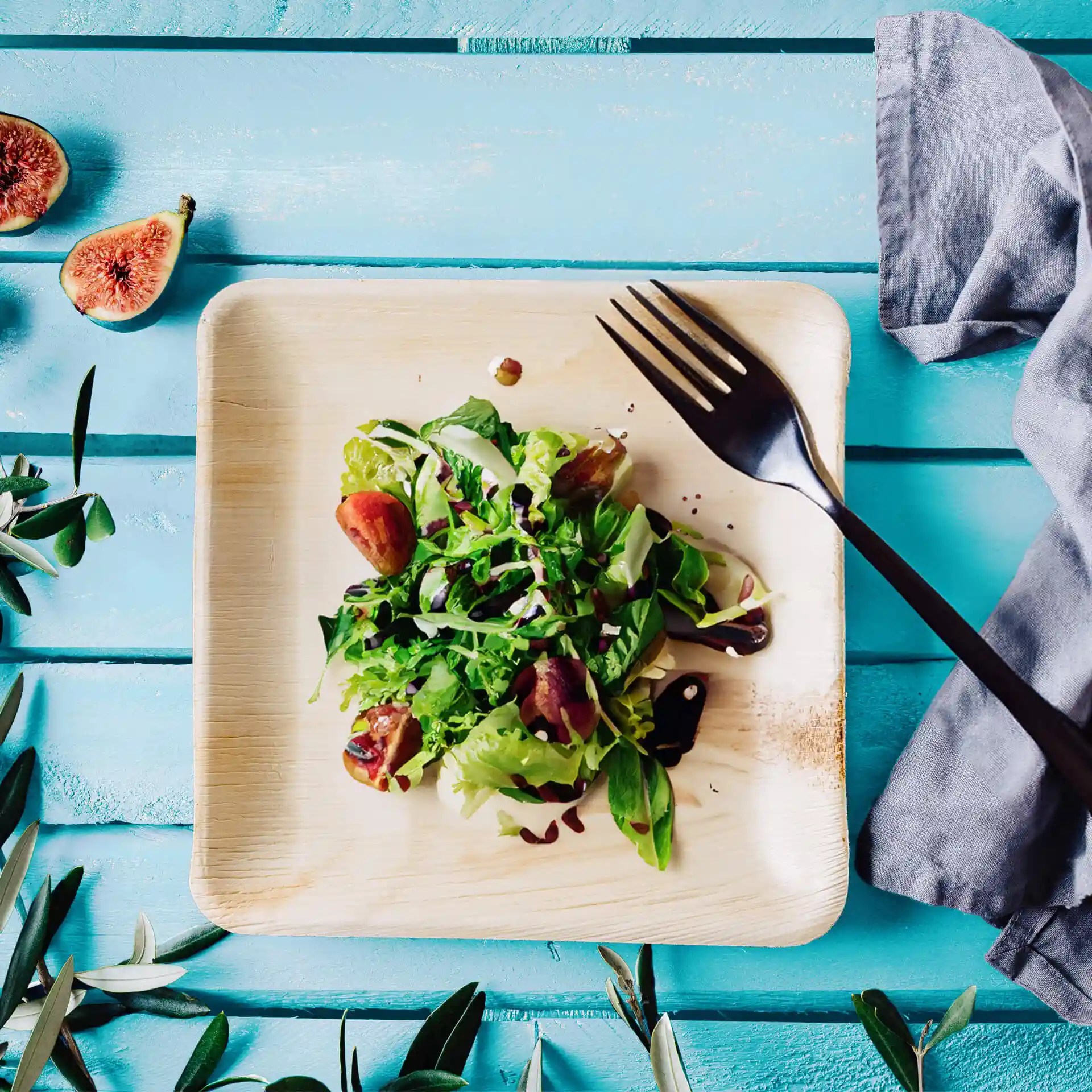 Assiette carrée en feuille de palmier avec une salade garnie de légumes verts et de figues, accompagnée d'une fourchette noire sur une surface en bois bleu.