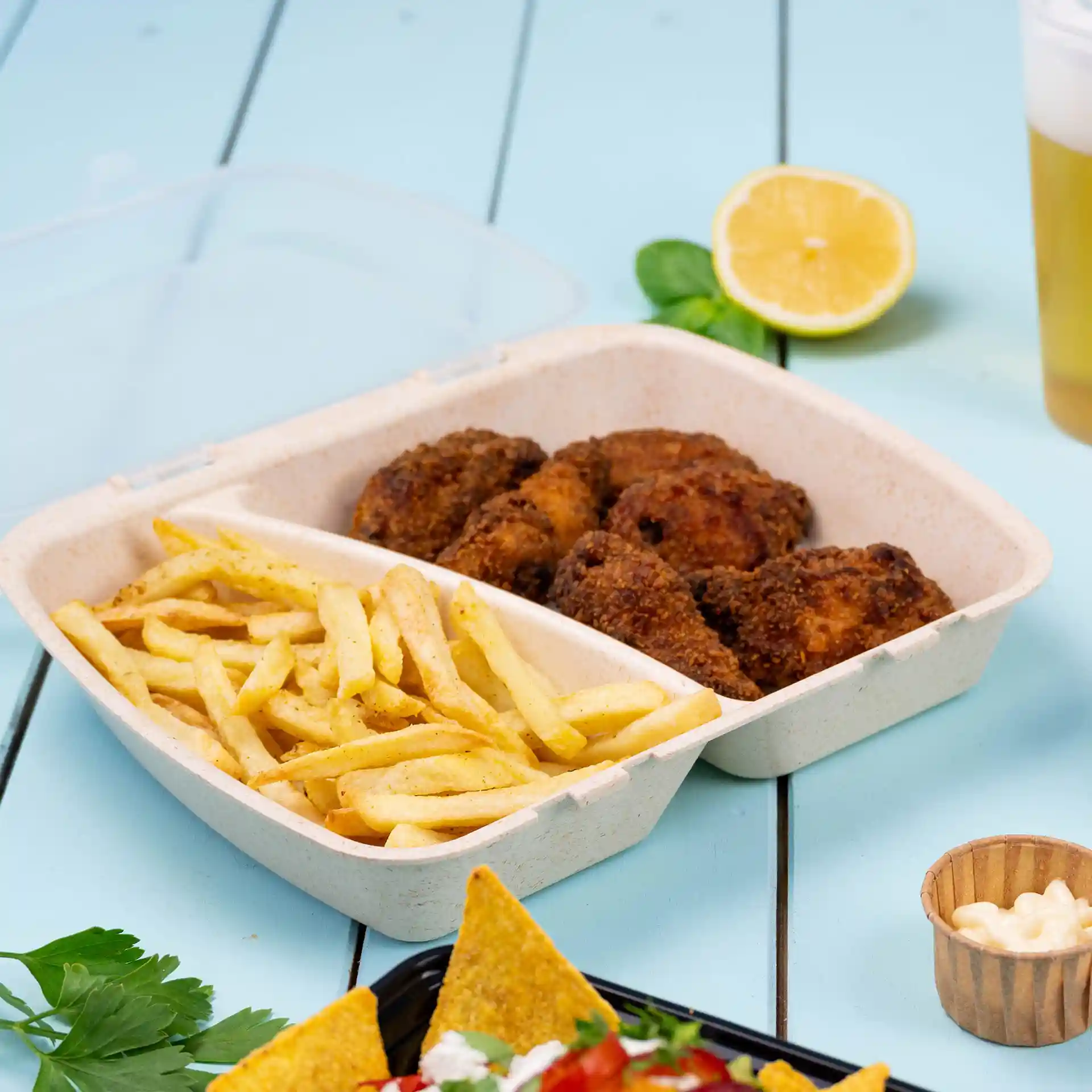 Conteneur à deux compartiments contenant du poulet frit et des frites, sur une surface bleue avec un citron et une boisson à proximité.