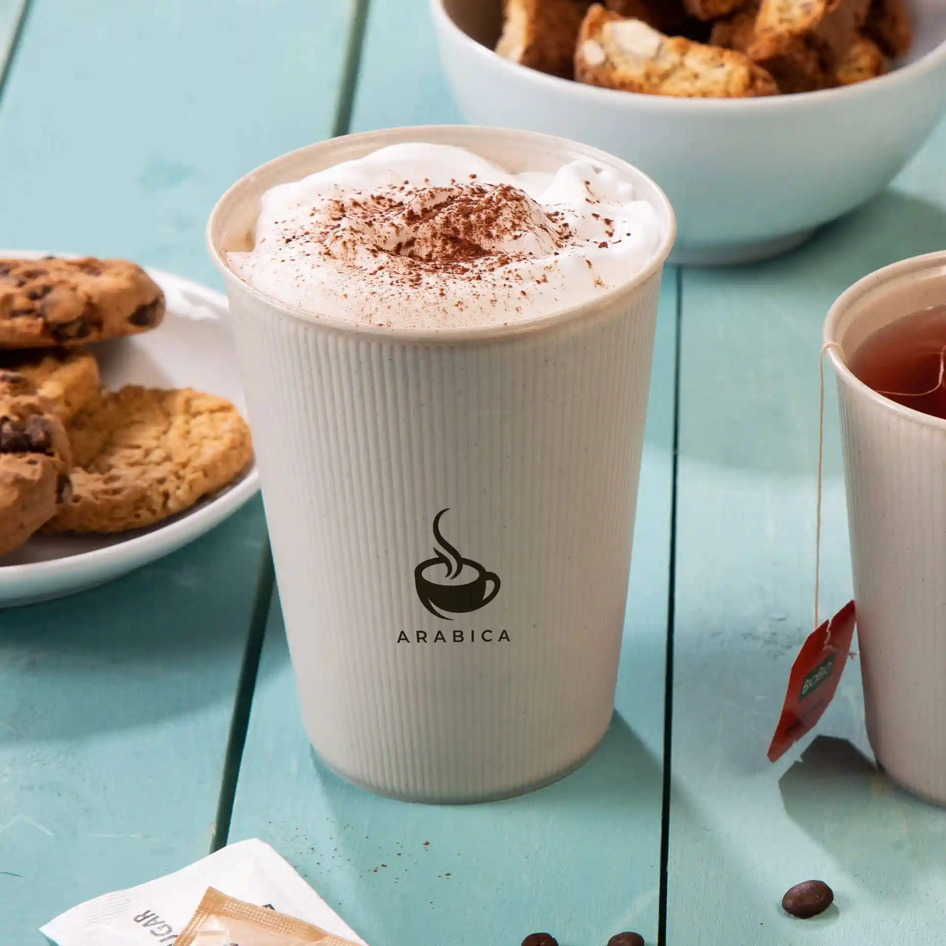 Gobelet réutilisable avec un design ondulé, rempli de crème fouettée et de poudre de cacao, à côté de biscuits et de thé.
