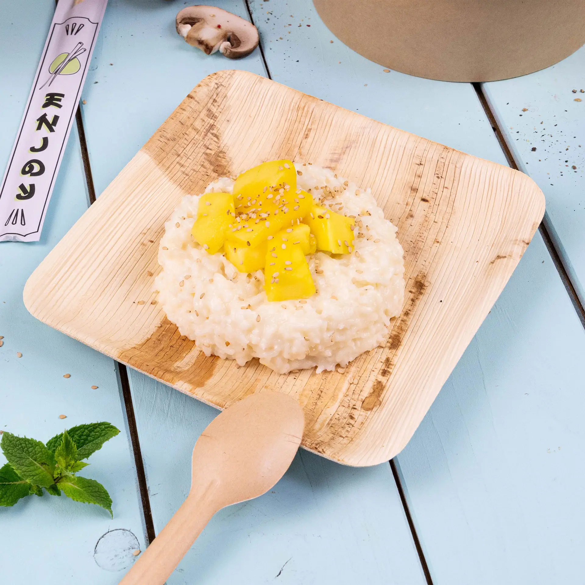 Assiette carrée en feuille de palmier avec risotto surmonté de mangue et de graines de sésame, accompagné d'une cuillère en bois et de feuilles de menthe.