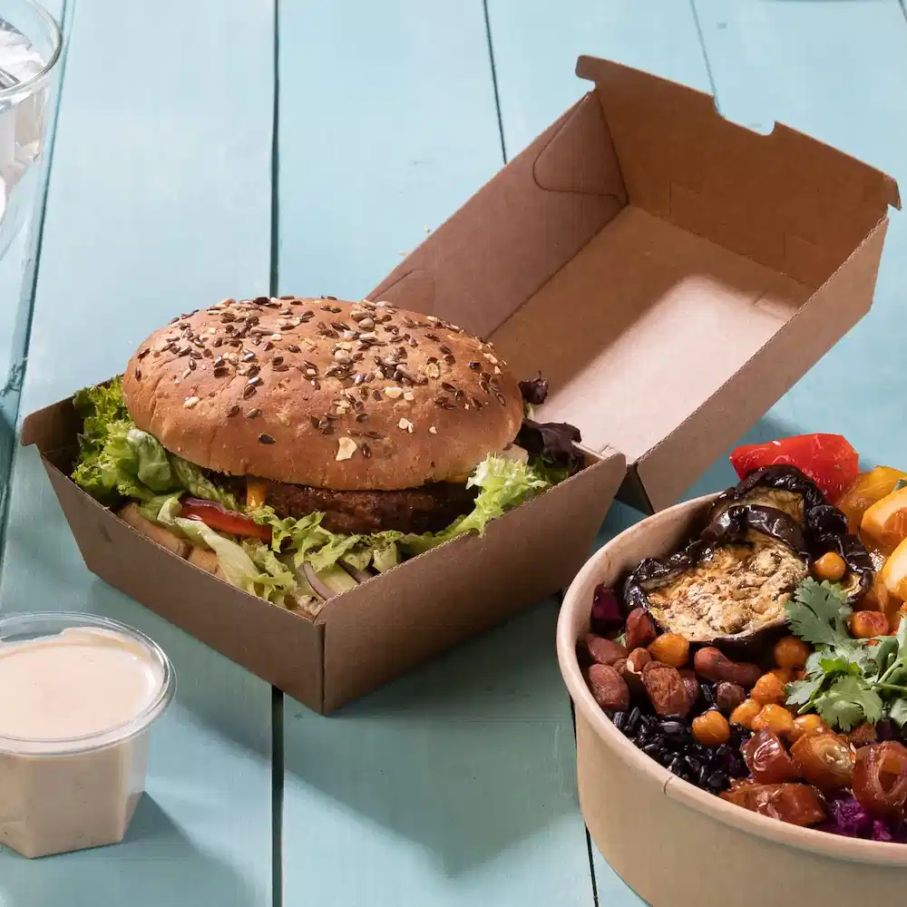 Un burger avec un pain aux graines de sésame et de la laitue dans une boîte à emporter brune, à côté d'un bol de légumes et légumineuses mélangés.