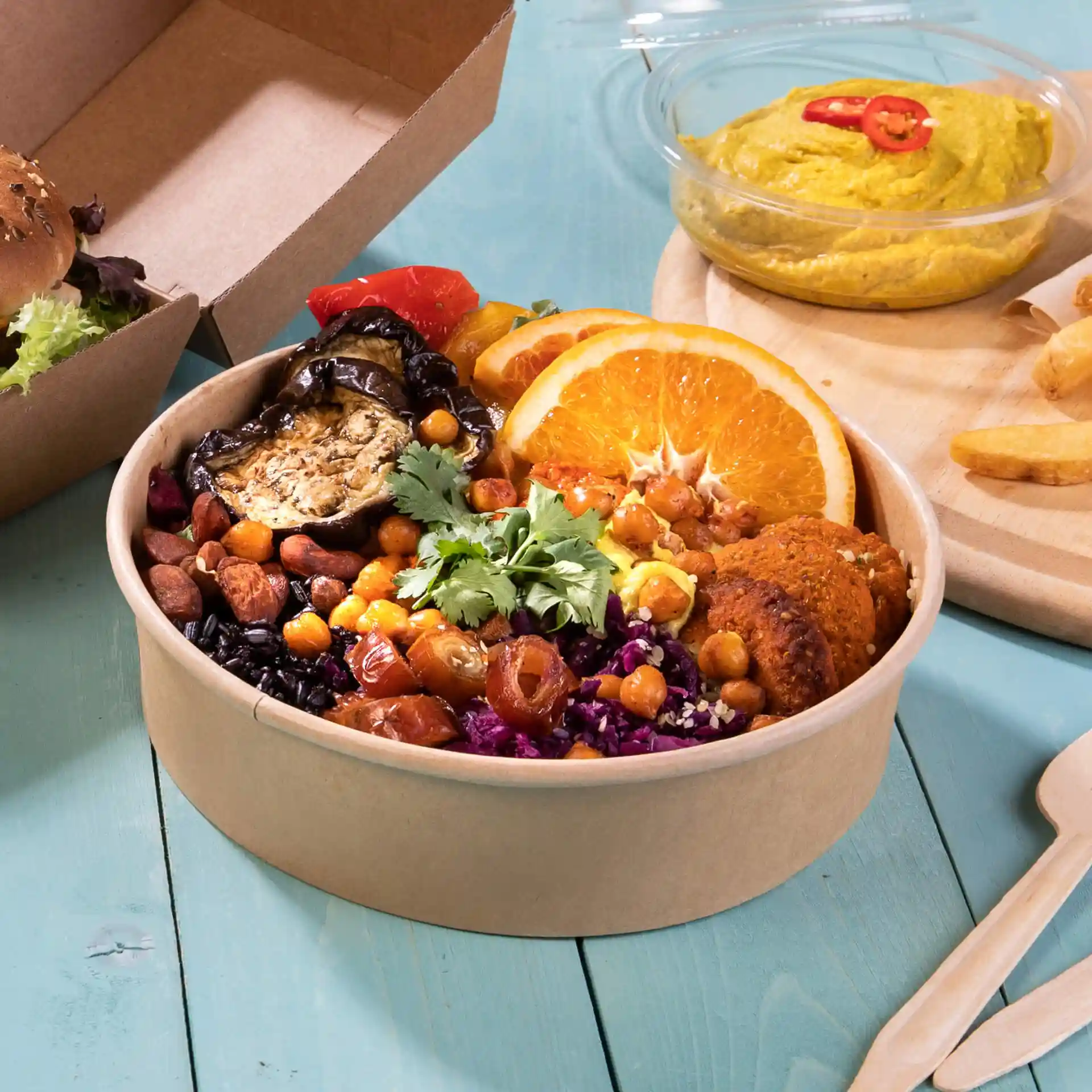 Bol rond en carton brun rempli de légumes variés, de pois chiches et de tranches d'orange, avec un accompagnement de houmous et de frites.