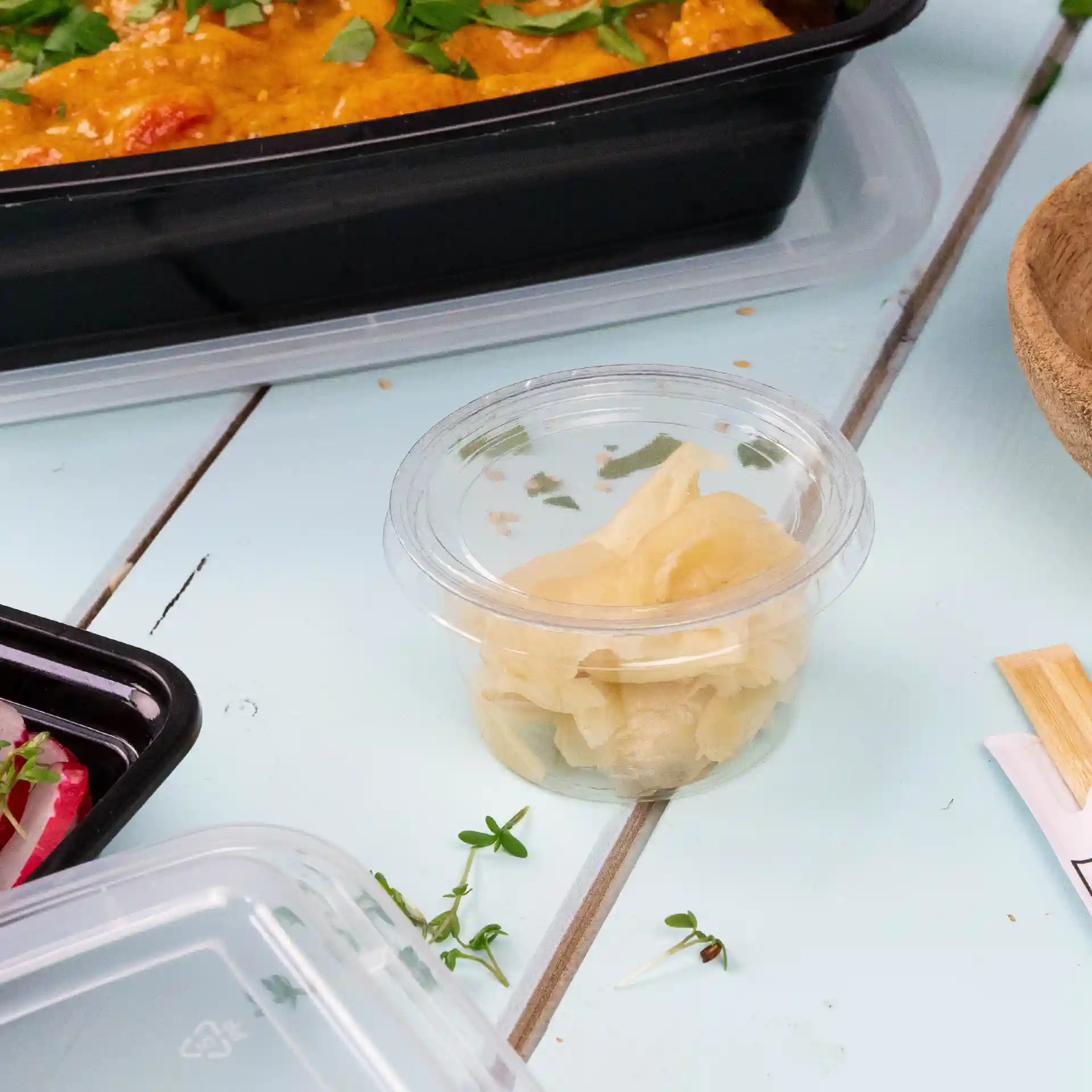 Gobelet en PLA transparent contenant du gingembre mariné, placé sur une surface bleu clair avec d'autres aliments à proximité.