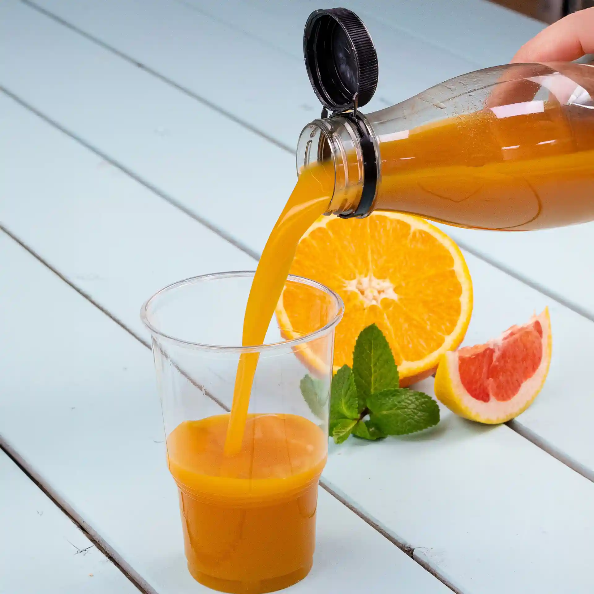 Bouteille rPET transparente versant du jus d'orange dans un verre transparent, avec des tranches d'orange et de pamplemousse à proximité.