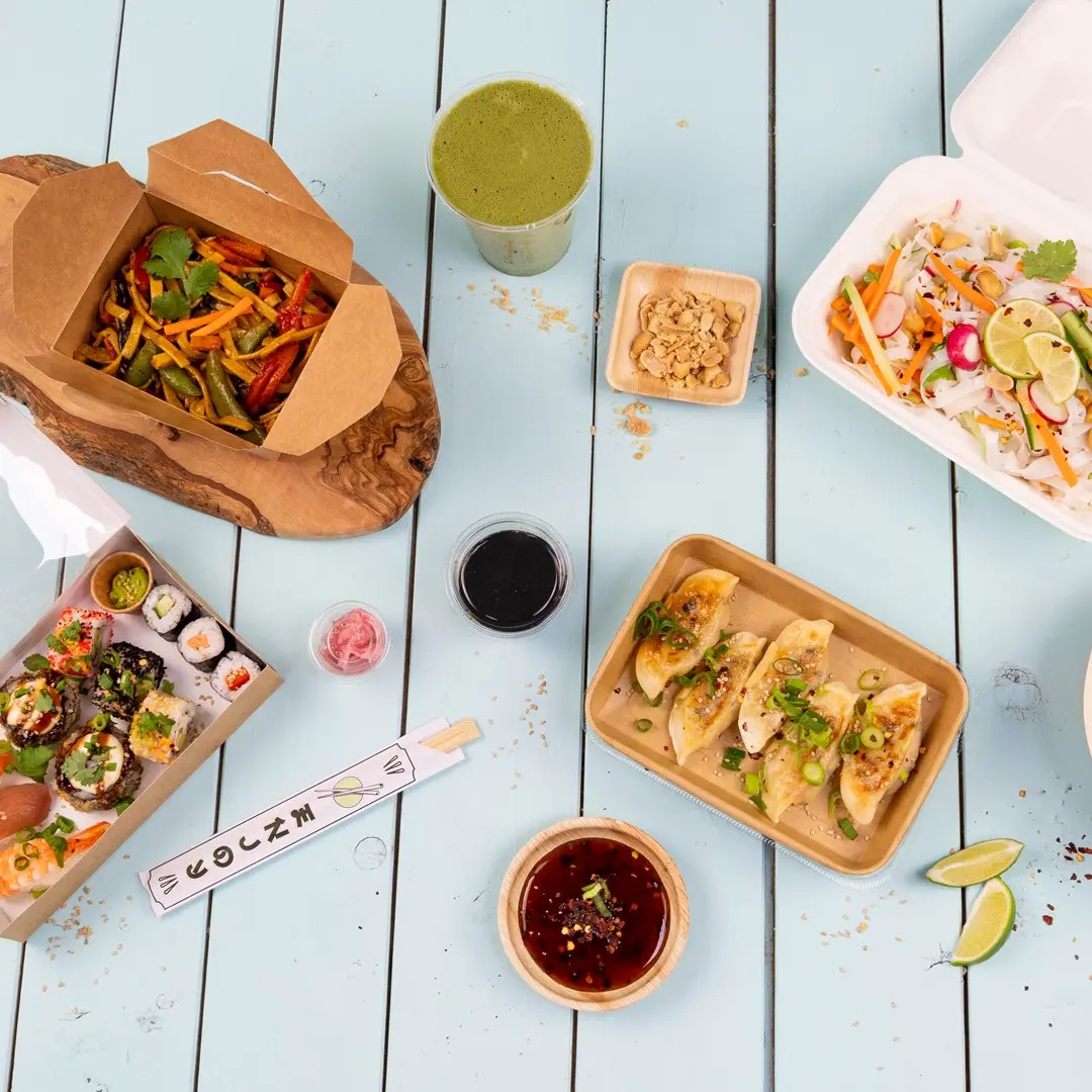 Assortiment de plats asiatiques comprenant des sushis, des dumplings, des légumes sautés et une boisson verte sur une table en bois bleue.