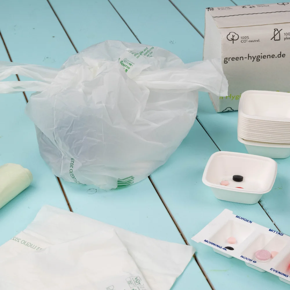 Sac en plastique contenant des produits d'hygiène; un bol blanc avec divers articles et une boîte étiquetée "green-hygiene.de" à proximité.