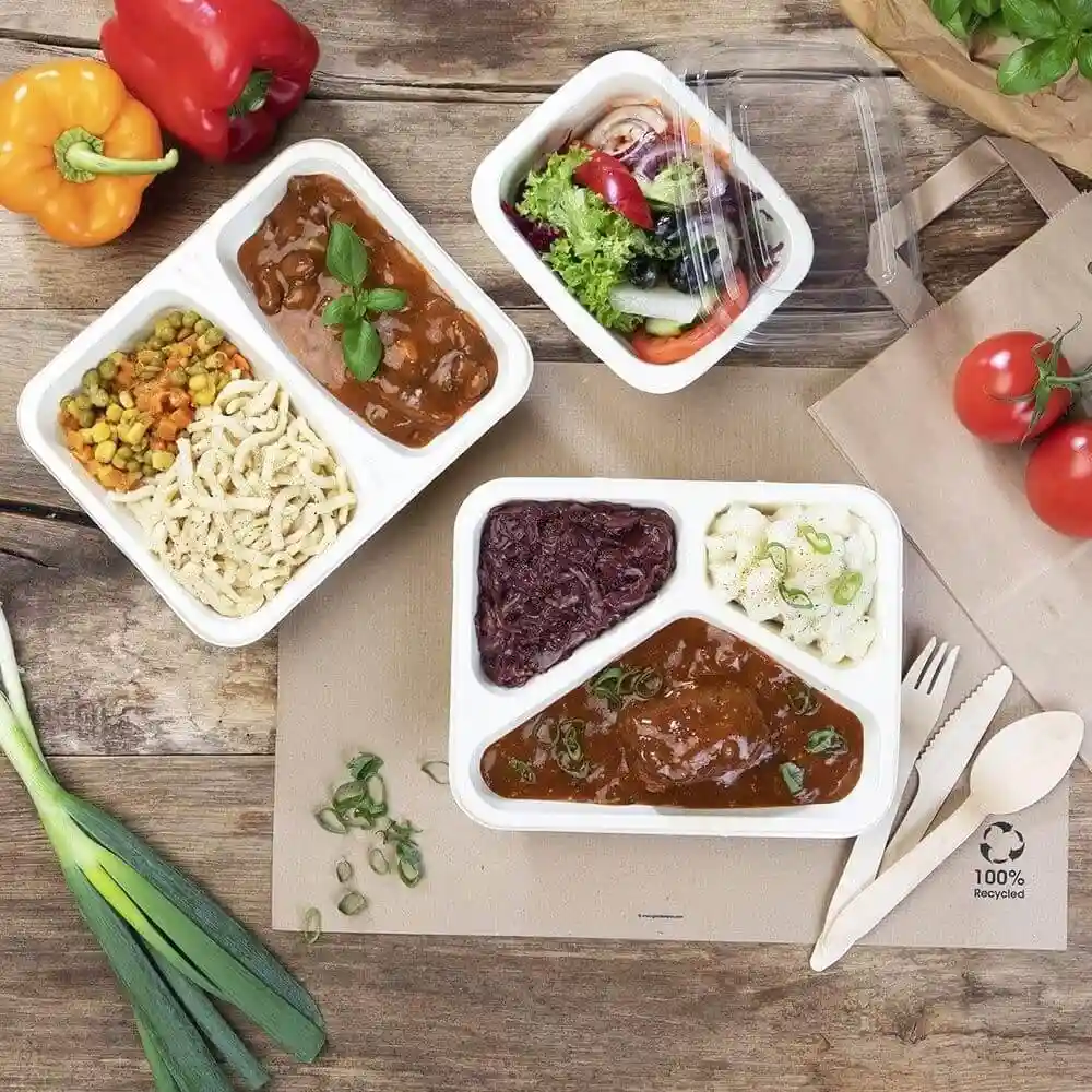 Image de deux plateaux en cellulose avec trois compartiments chacun, contenant divers aliments, sur une surface en bois avec des légumes.