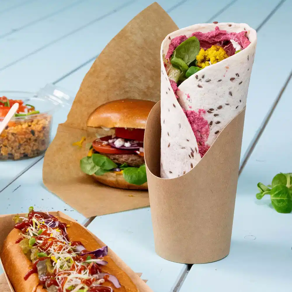 Un wrap plié aux légumes est présenté dans un support en papier, avec un burger et un hot-dog à proximité.