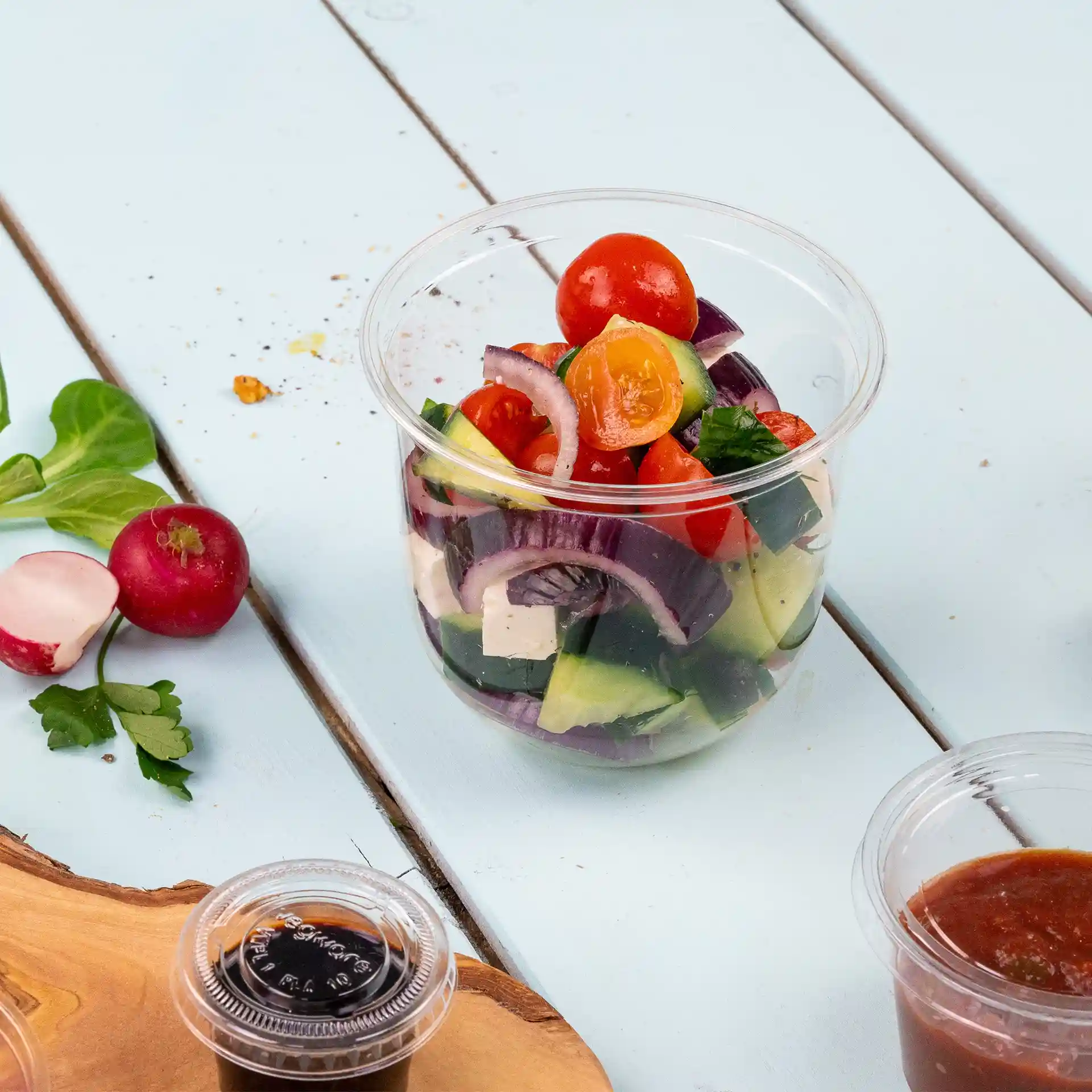 Gobelet transparent rempli d'ingrédients de salade mélangés, y compris des tomates, du concombre et des oignons, sur une surface bleu clair.