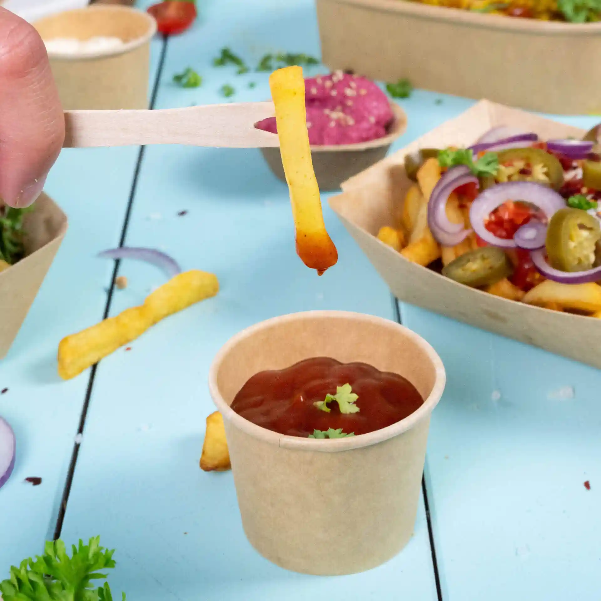 Image d'une main tenant une fourchette en bois avec une frite trempée dans du ketchup au-dessus d'un petit gobelet marron de ketchup.