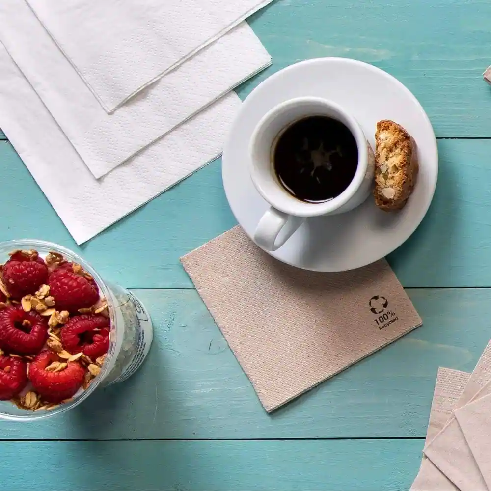 Tasse de café sur une soucoupe avec un biscotti, à côté d'un contenant transparent de framboises et de granola, sur une surface en bois bleue.
