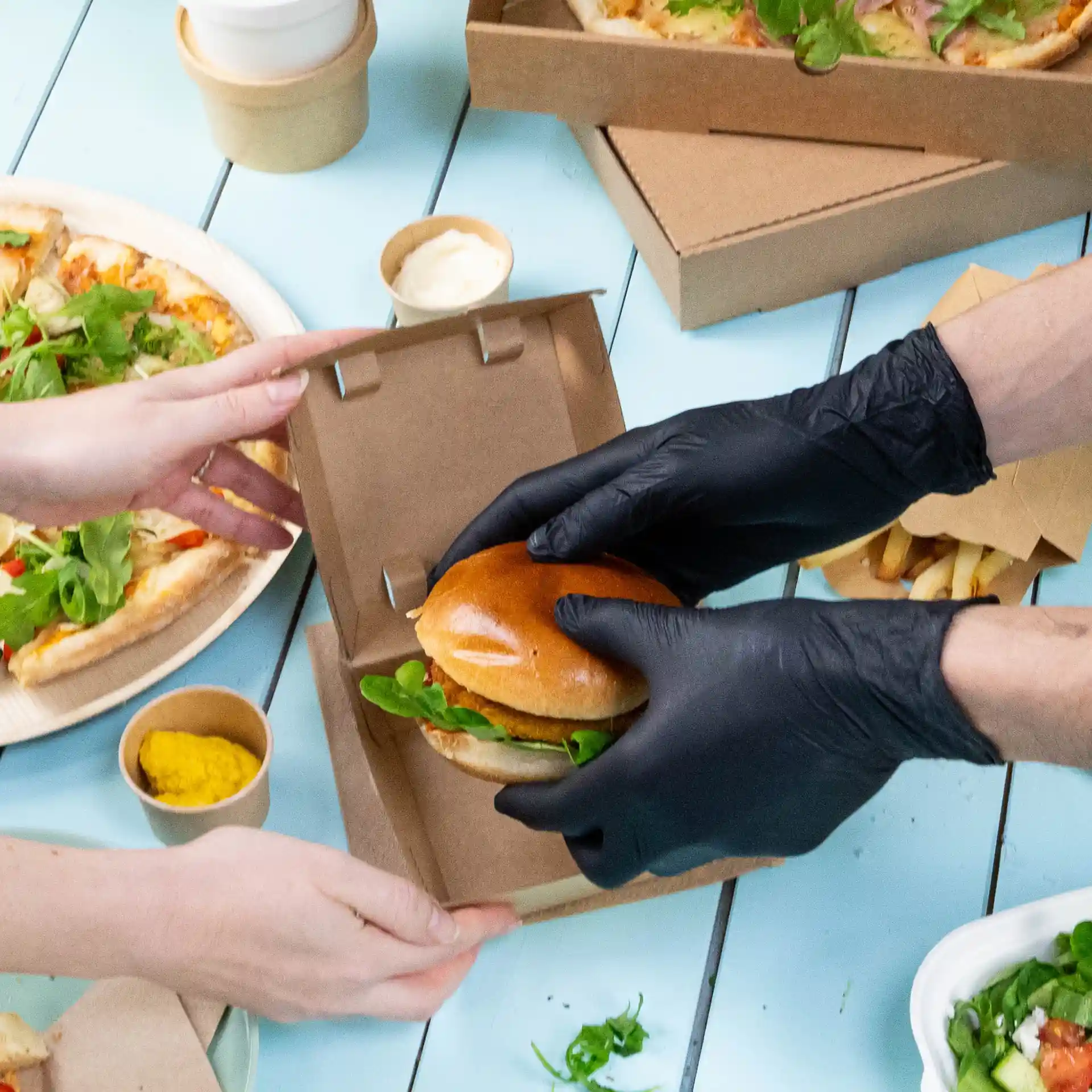 Image d'une personne portant des gants en nitrile noirs remettant un burger à une autre personne, avec de la pizza et des condiments en arrière-plan.