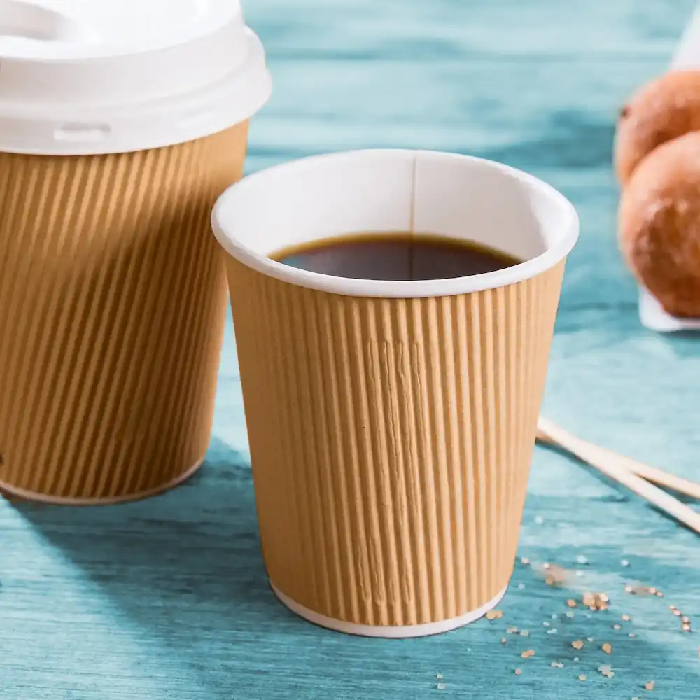 Gobelet en papier brun ribbé avec du café noir, à côté d'un gobelet similaire et de donuts sur une surface bleue.