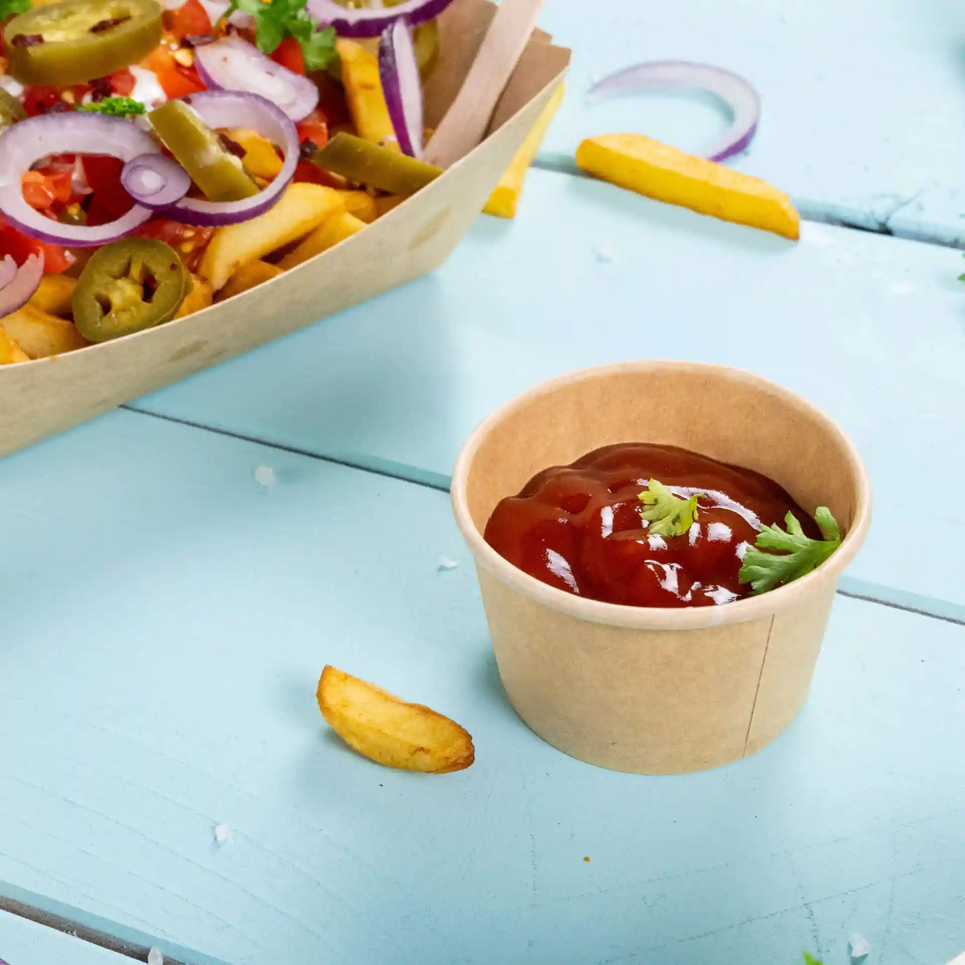 Gobelet en papier brun avec du ketchup et une garniture de coriandre, posé sur une surface bleu clair.