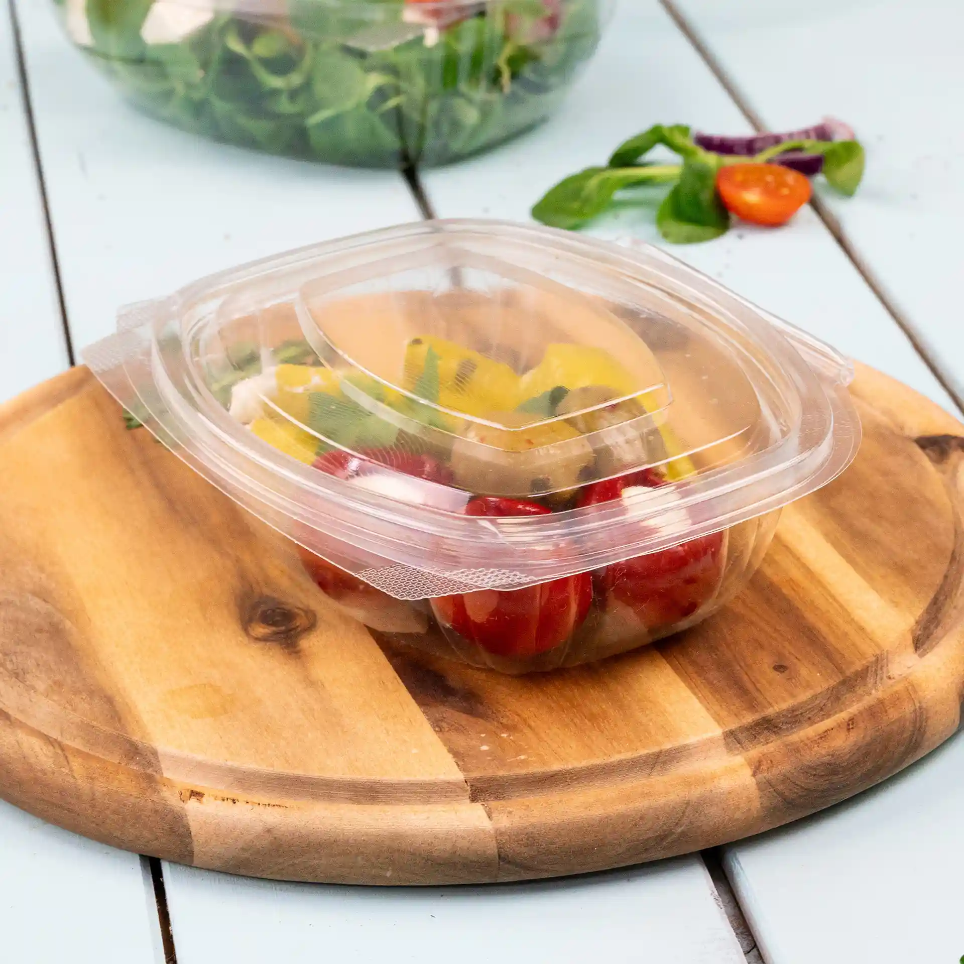 Bol rectangulaire en PLA transparent avec couvercle pliant, contenant des légumes mélangés, posé sur une planche à découper en bois.