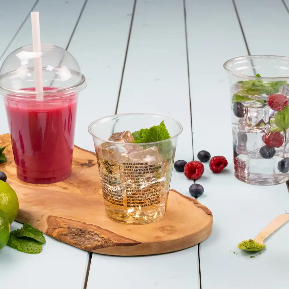 Des gobelets en plastique transparent avec des boissons et de la menthe posés sur une planche en bois, avec des baies et du citron vert éparpillés.