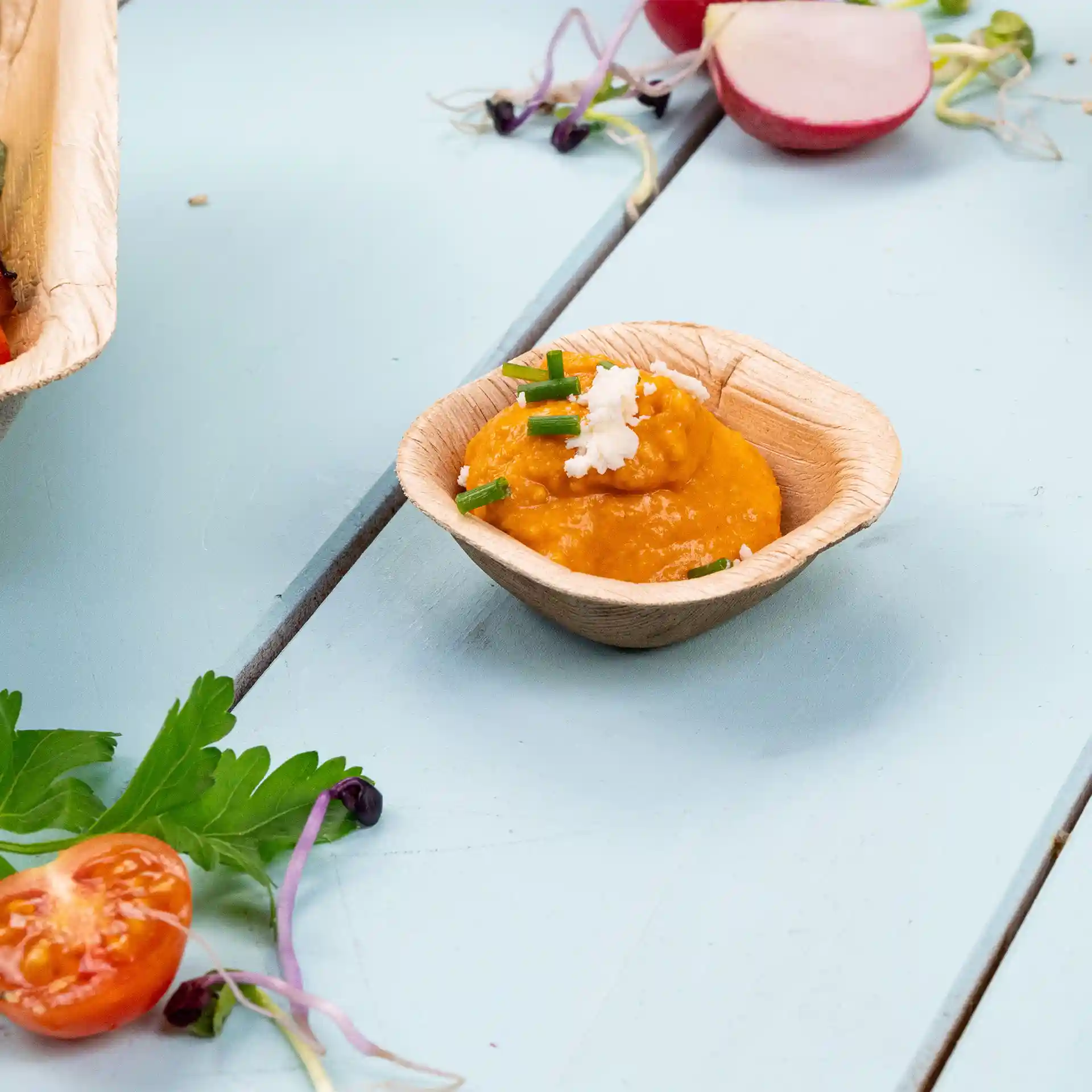 Bol carré profond en feuille de palmier contenant une sauce orange garnie de ciboulette et de fromage, posé sur une surface bleu clair.