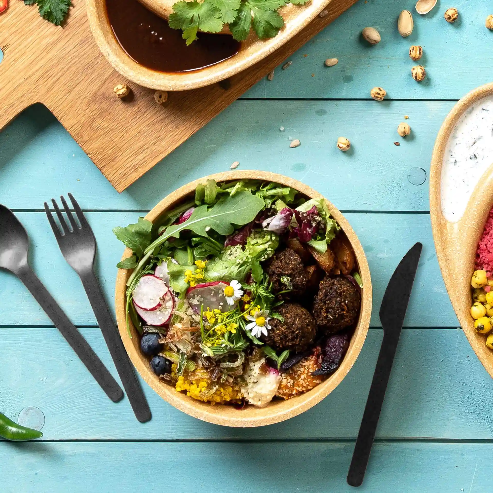 Bol avec salade, falafels et fleurs comestibles, accompagné de couverts réutilisables noirs sur une surface en bois bleu.