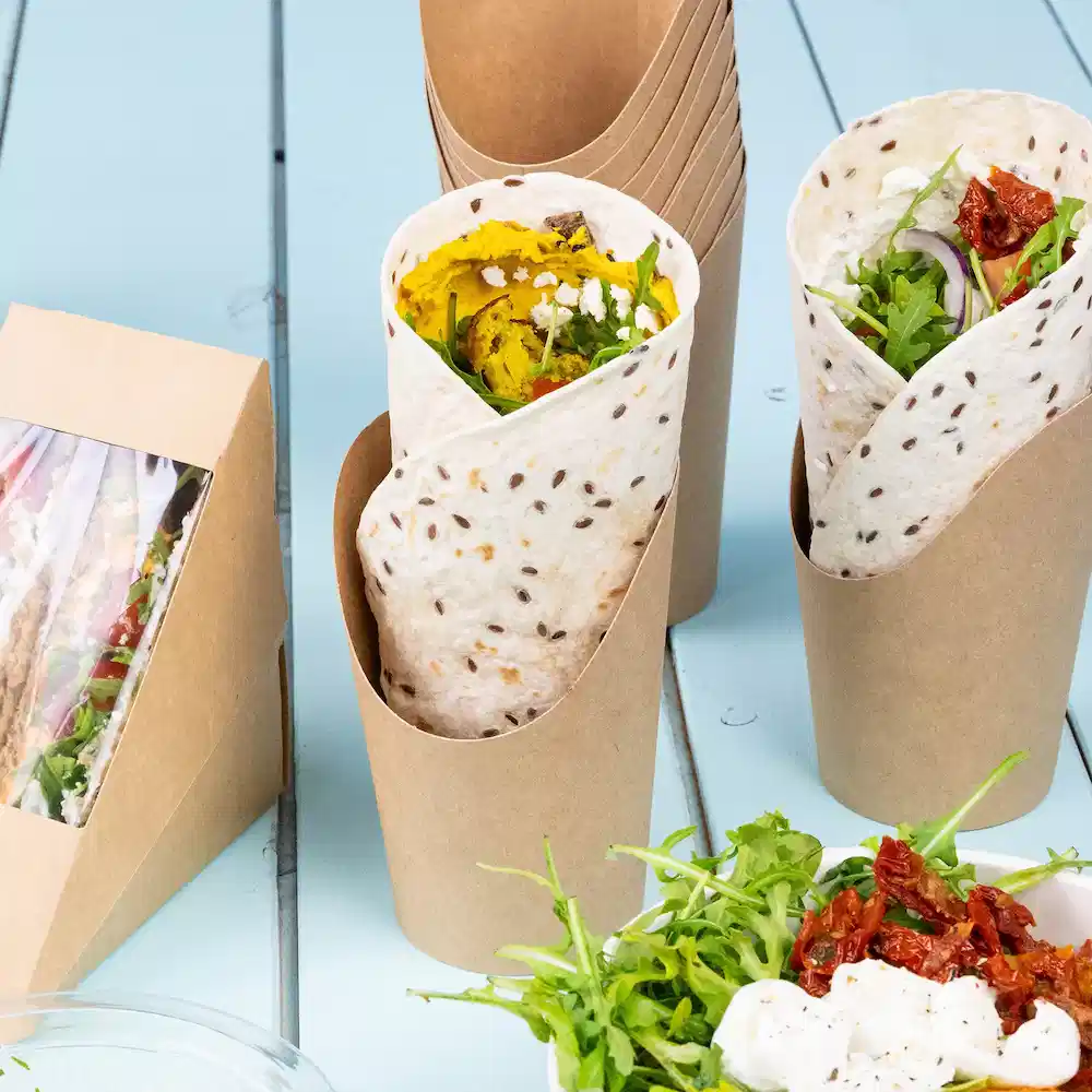 Deux wraps dans des récipients en papier avec les ingrédients visibles, accompagnés d'une salade et de tomates séchées sur une table.