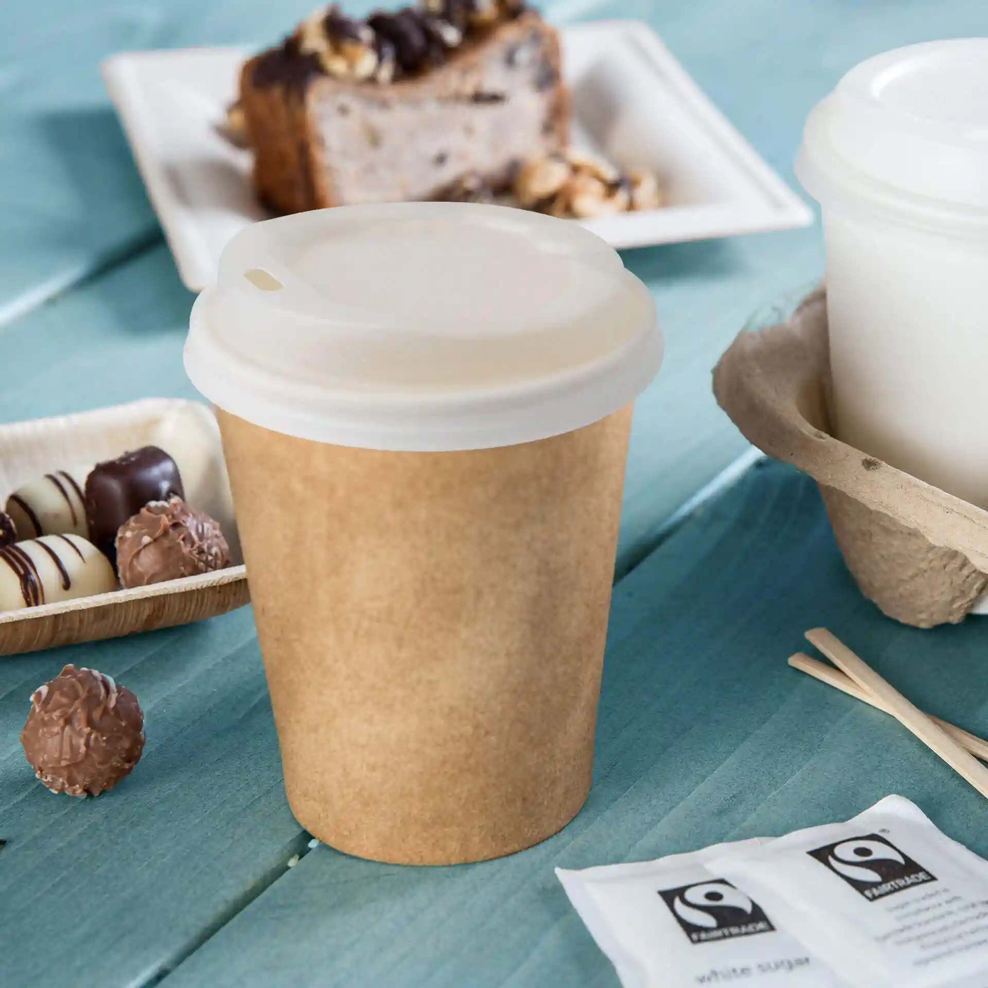 Gobelet en papier brun avec un couvercle blanc, posé sur une surface en bois bleu, avec des chocolats et des sachets de sucre à proximité.