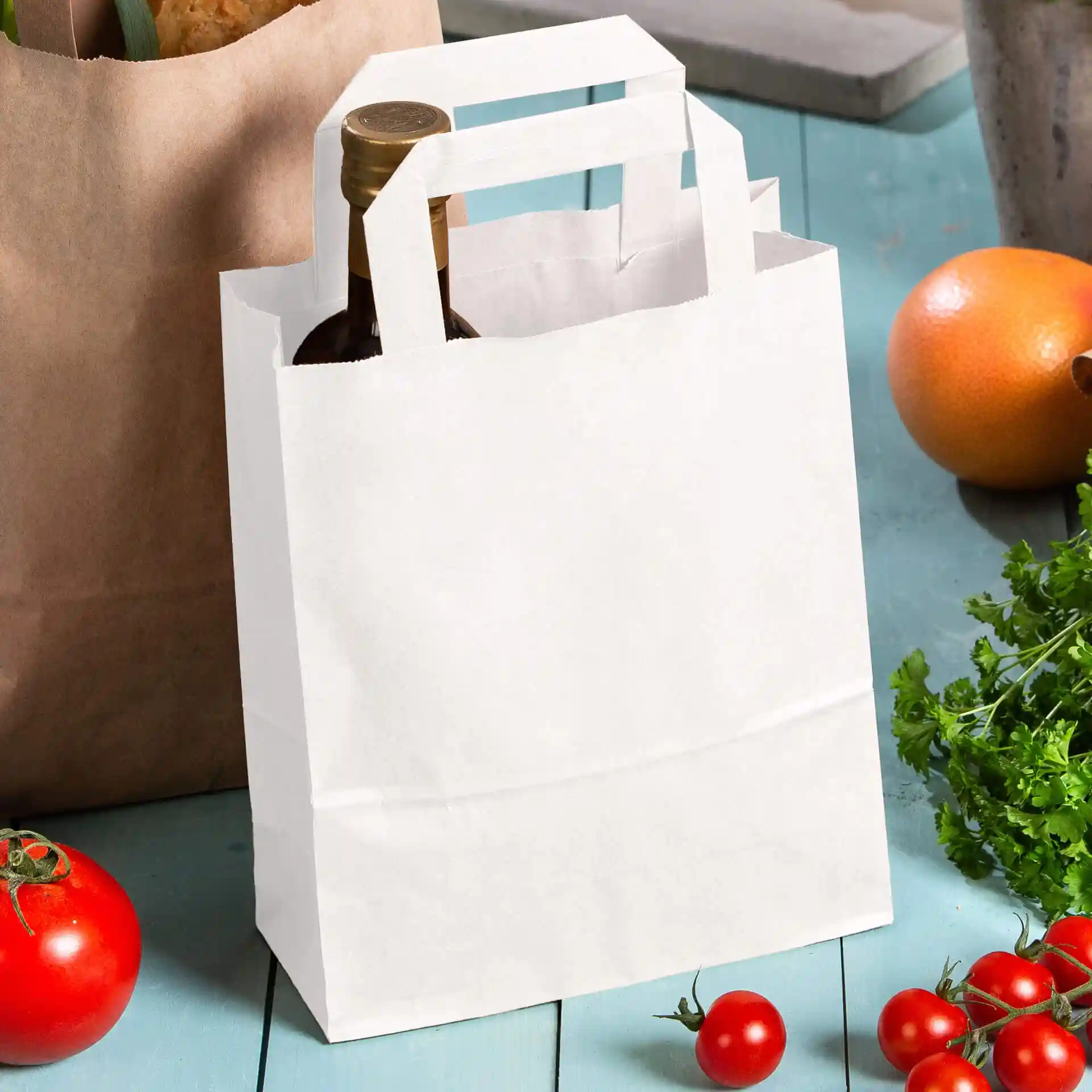 Sac en papier kraft blanc avec poignées, tenant une bouteille, entouré de tomates et d'une orange sur une surface bleue.