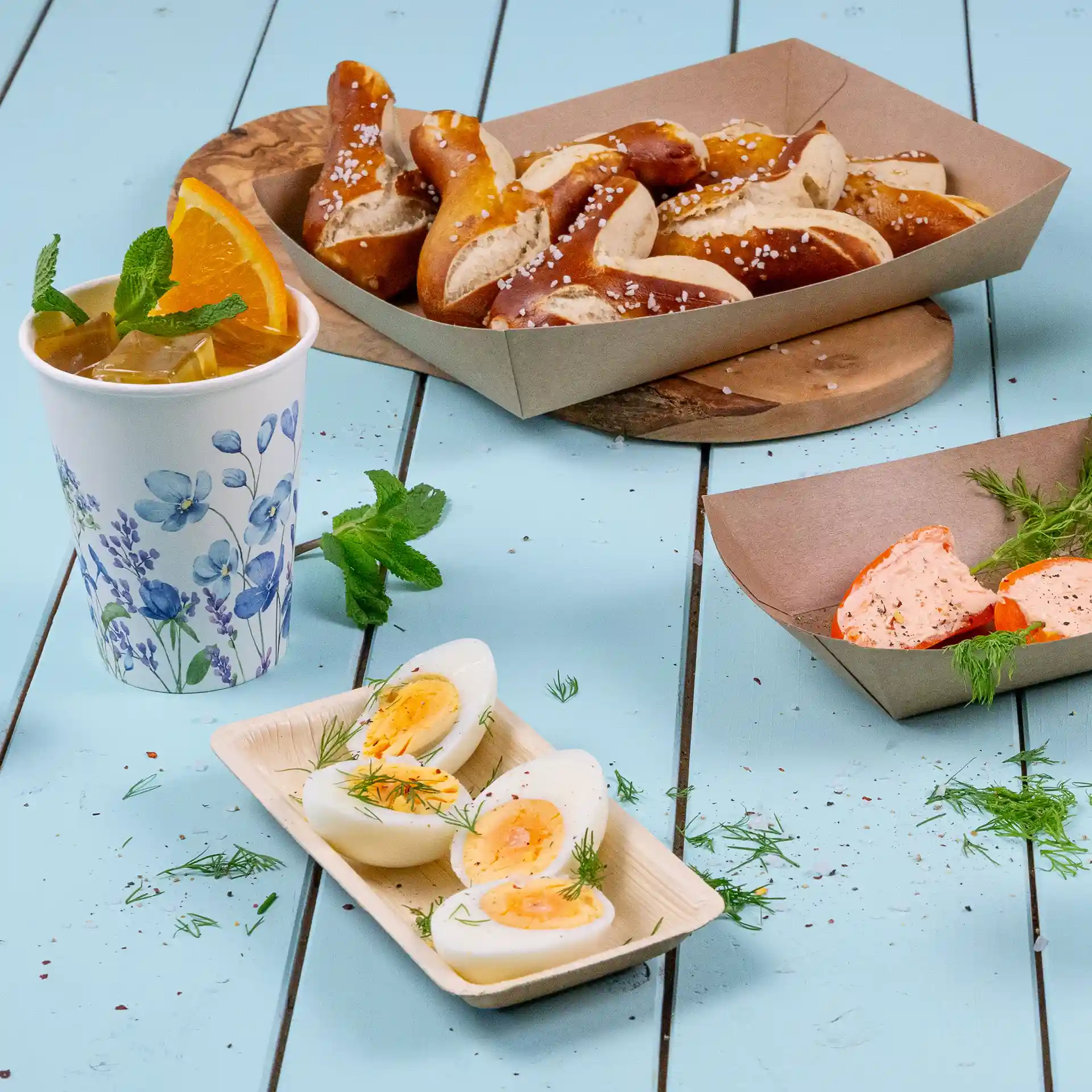 Image d'un verre avec un design floral, des bretzels dans une boîte brune et des œufs durs tranchés sur un plateau en feuille de palmier.