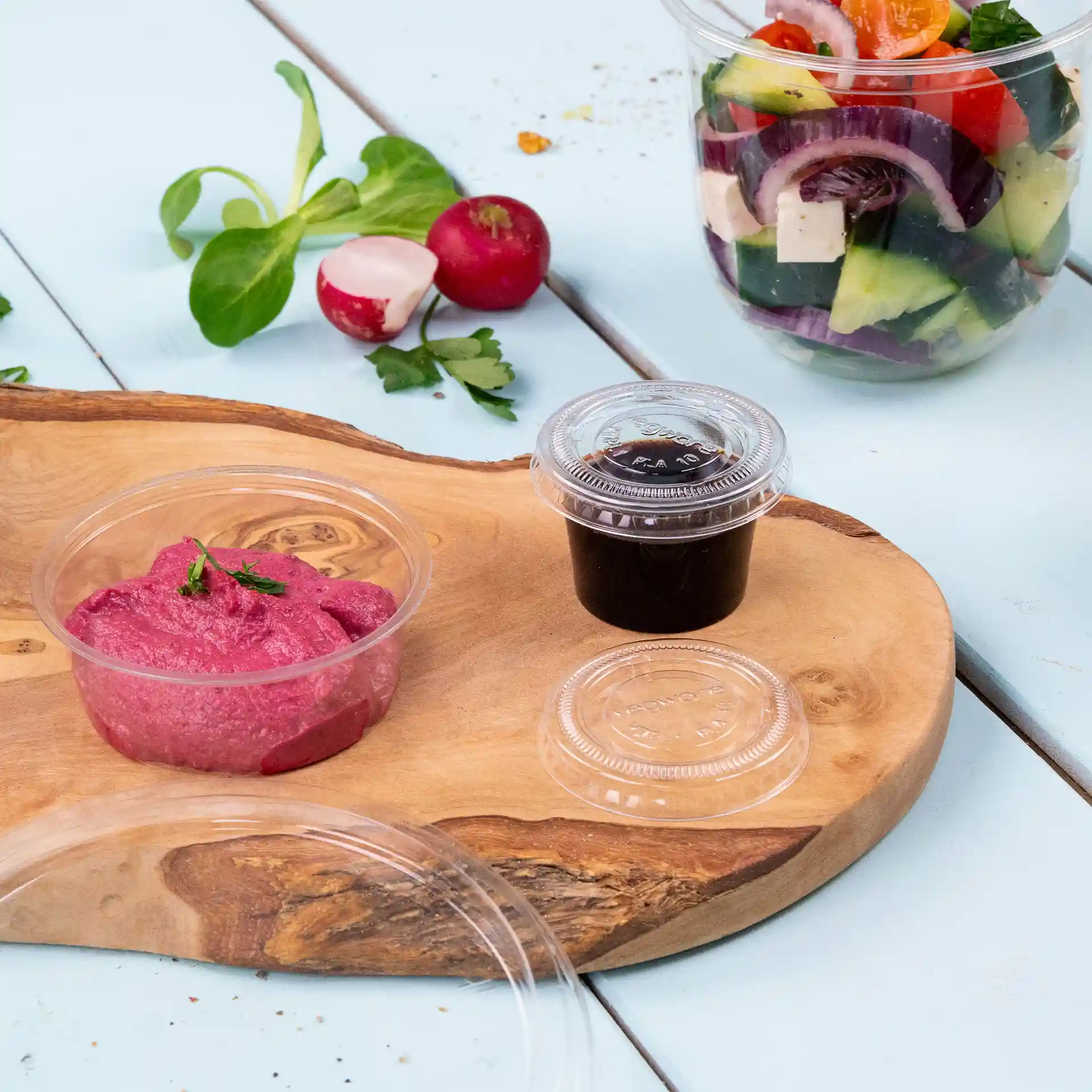 Coupes transparentes avec sauce rose et sauce foncée sur une planche en bois, avec salade et légumes en arrière-plan.