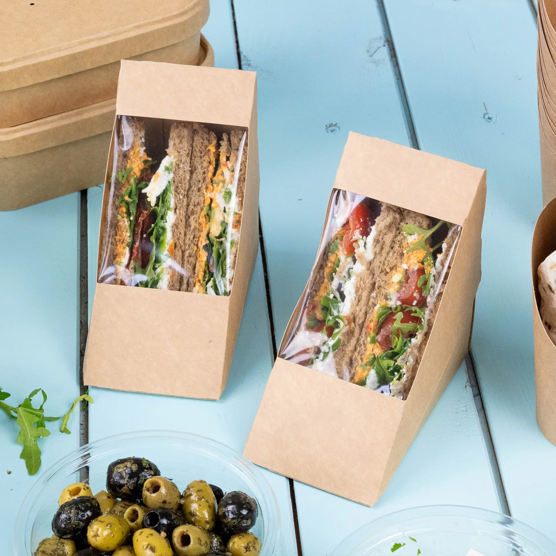 Deux coins de sandwichs en carton brun avec une fenêtre en PLA transparent montrant des sandwichs et des petits pains sur une surface bleue, avec des olives dans un bol à proximité.