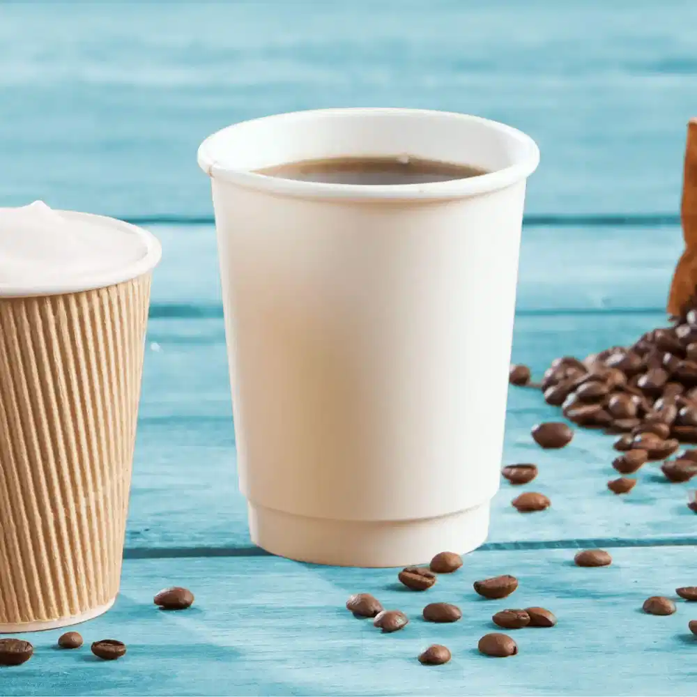 Deux gobelets en papier à double paroi empilés, un blanc uni et un en carton brun, avec des grains de café éparpillés.