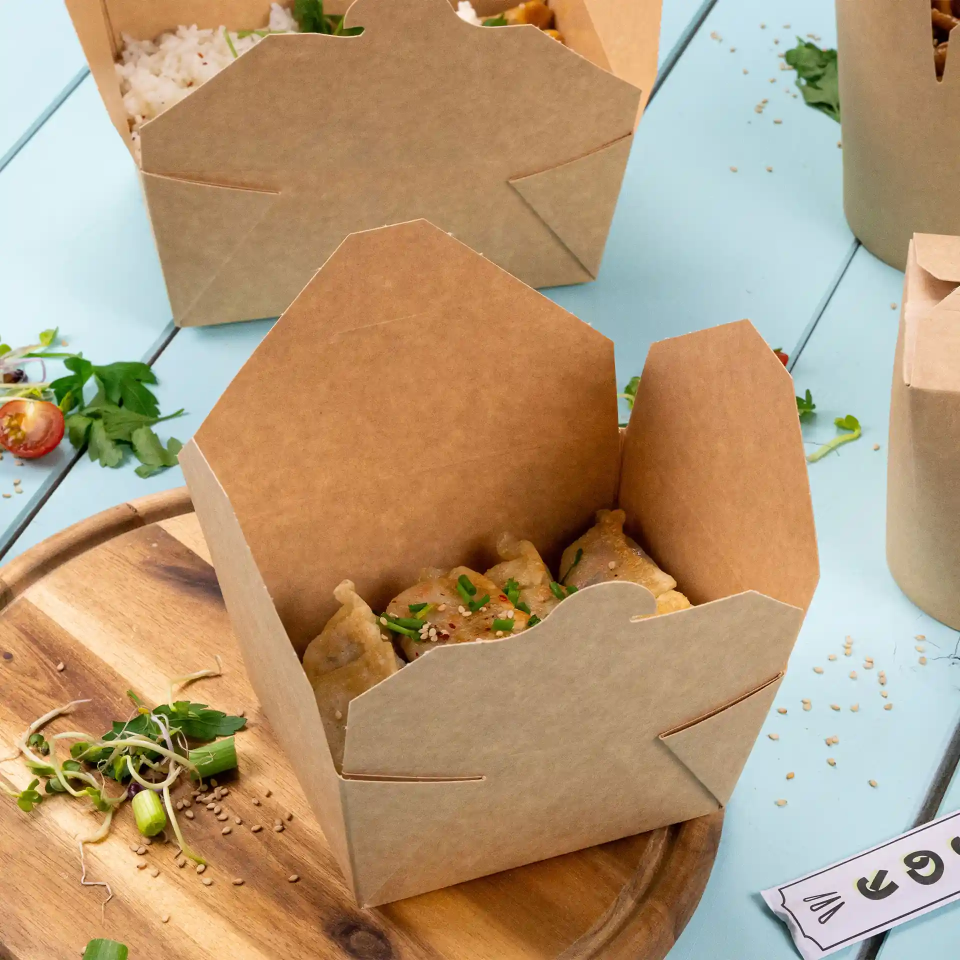 Boîte à emporter en carton brun avec des dumplings à l'intérieur, placée sur une surface en bois avec des graines de sésame et des légumes éparpillés.