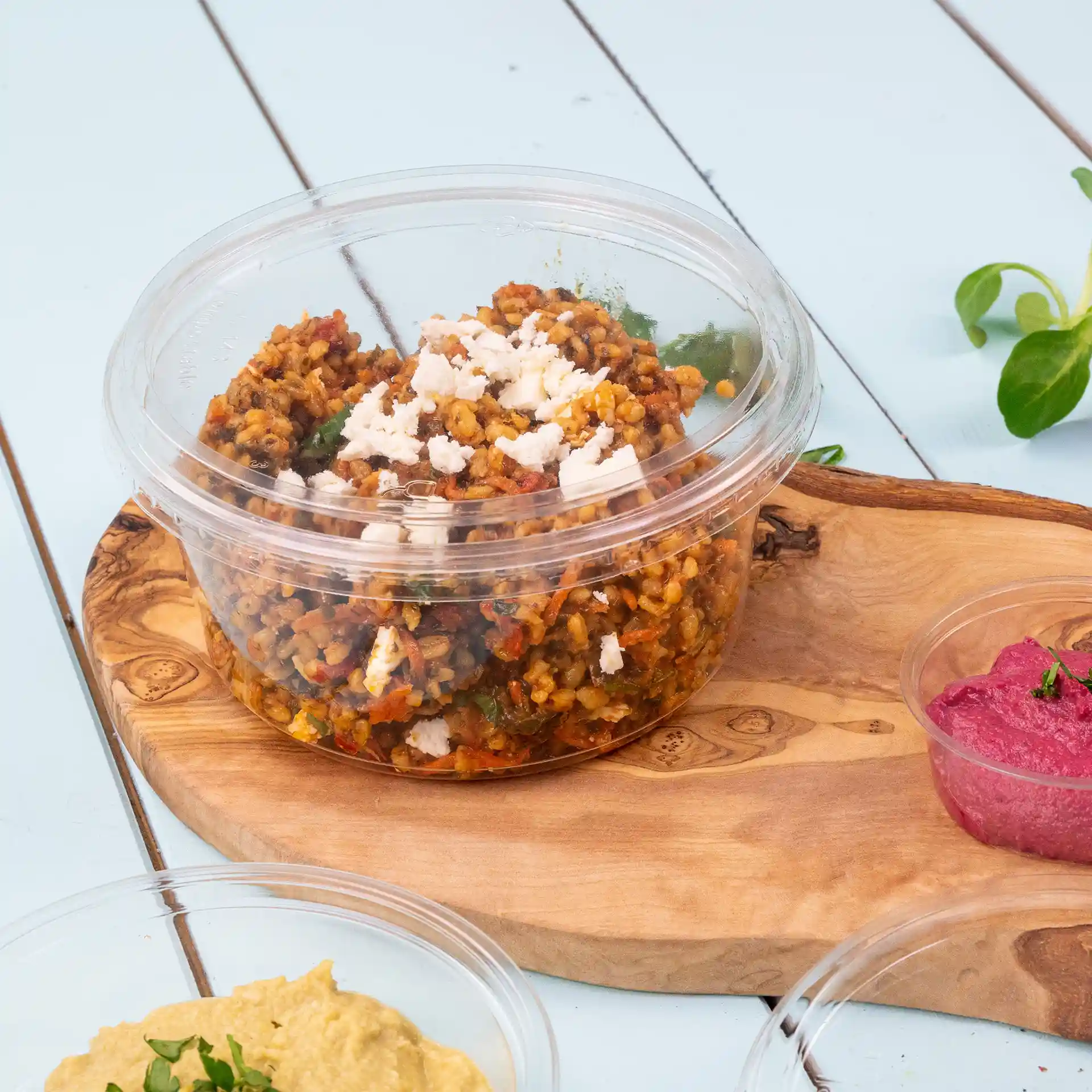 Gobelet rond transparent avec couvercle contenant un plat de céréales surmonté de fromage feta, posé sur une planche en bois.