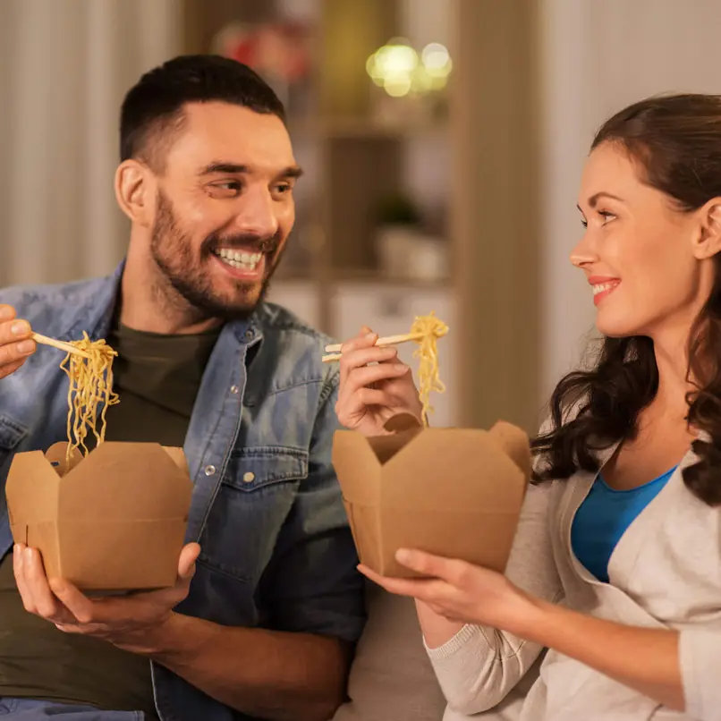Couple dégustant des nouilles à emporter dans des boîtes brunes, utilisant des baguettes, assis ensemble dans un cadre intérieur confortable.