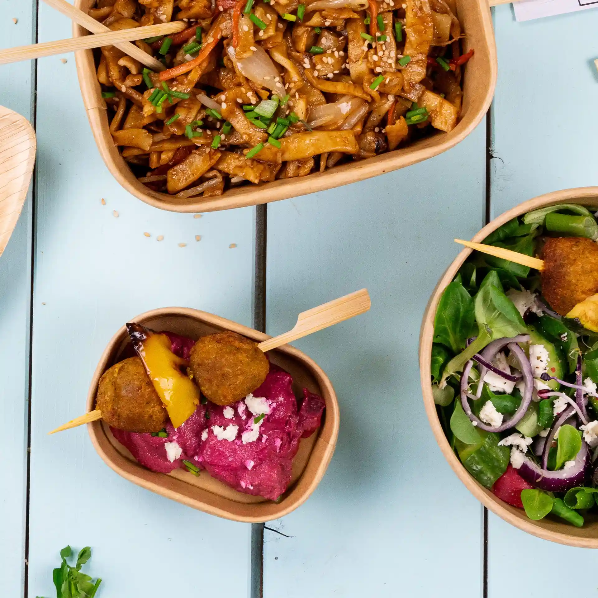 Image de trois contenants alimentaires en kraft rectangulaires avec divers plats : nouilles, salade et un dip de betterave avec des brochettes.