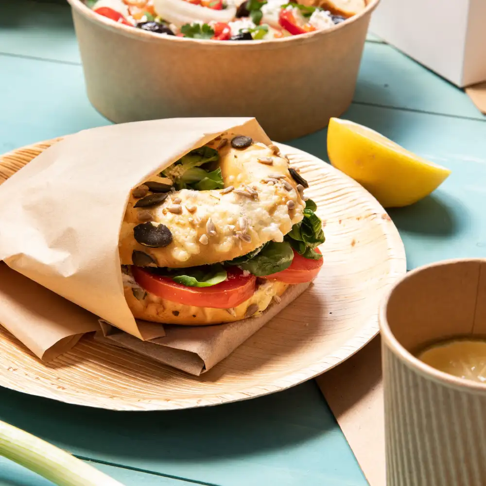 Sandwich bagel avec laitue et tomate sur une assiette en bois; un quartier de citron et une tasse sont également visibles en arrière-plan.