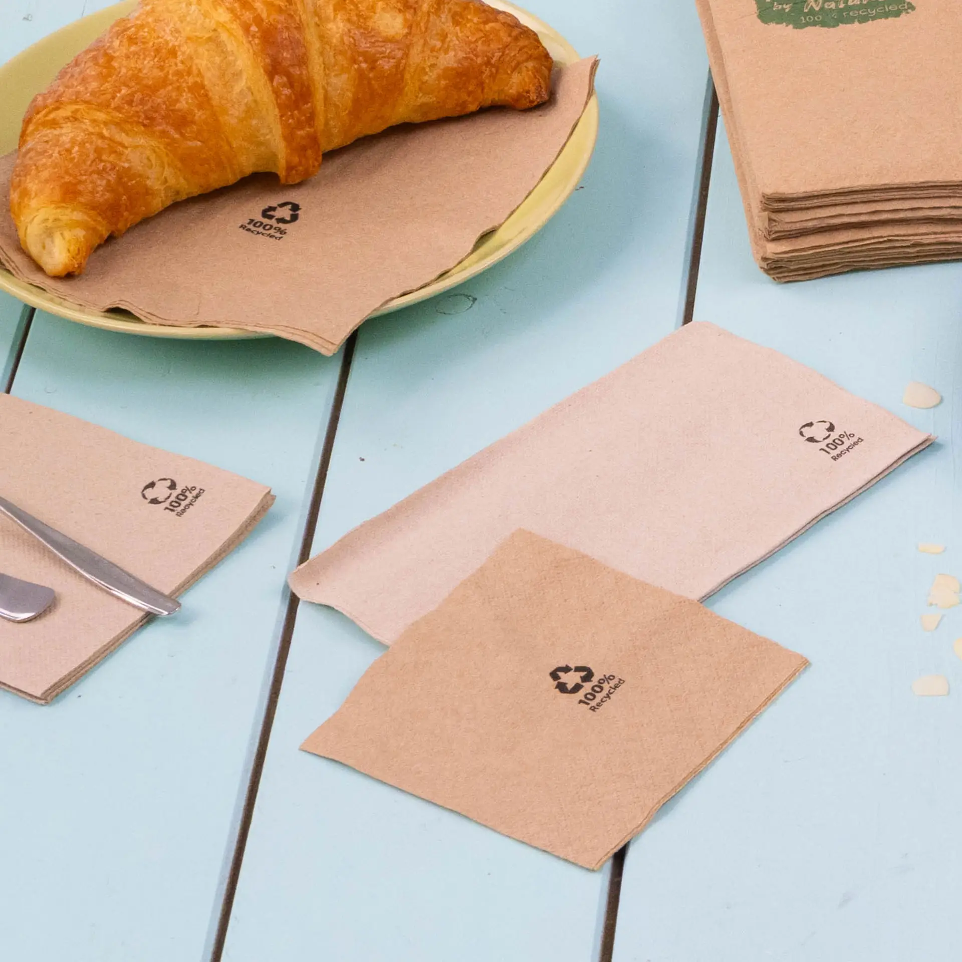 Image de serviettes à cocktail en papier recyclé marron, avec des logos de recyclage subtils, posées sur une table près d'un croissant et des couverts.