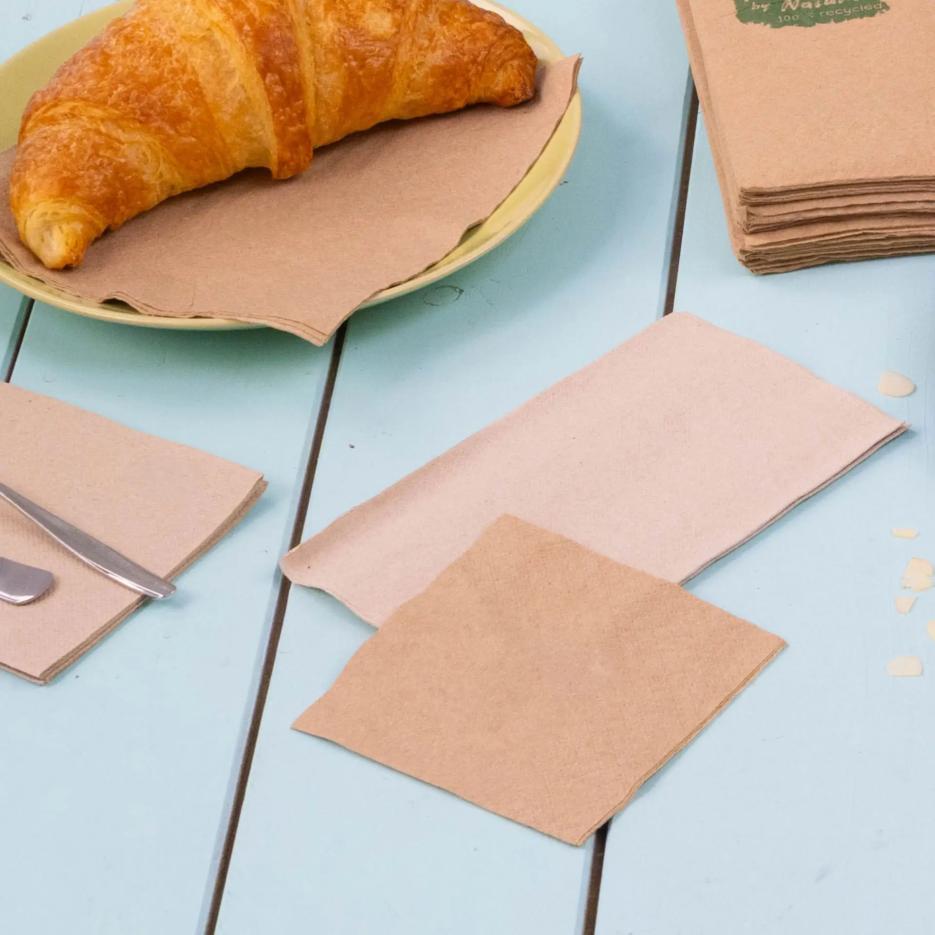 Image d'un croissant sur une assiette jaune avec plusieurs serviettes à cocktail en papier recyclé marron disposées sur une table bleue.