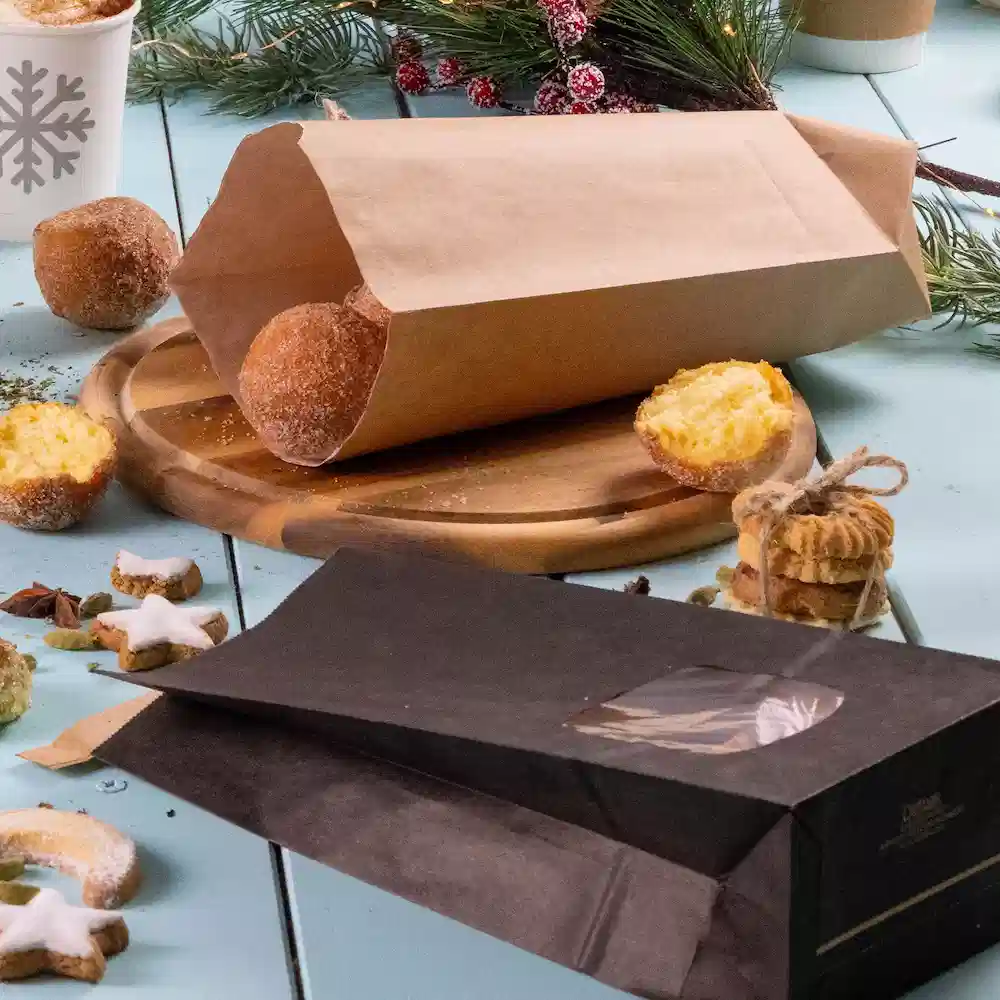 Sac en papier brun avec des produits de boulangerie, sac noir avec une fenêtre transparente, planche de service en bois et décorations festives.