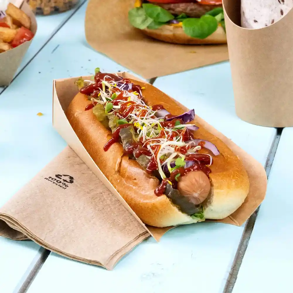 Hot-dog dans un support en papier brun, garni de ketchup, moutarde, cornichons et pousses, reposant sur une table en bois.