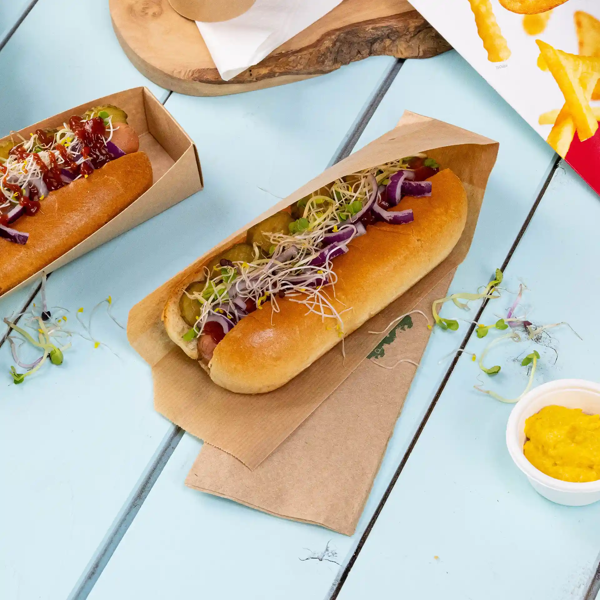Deux hot-dogs dans des plateaux en papier brun, garnis de divers légumes et sauces, sur une surface bleu clair.