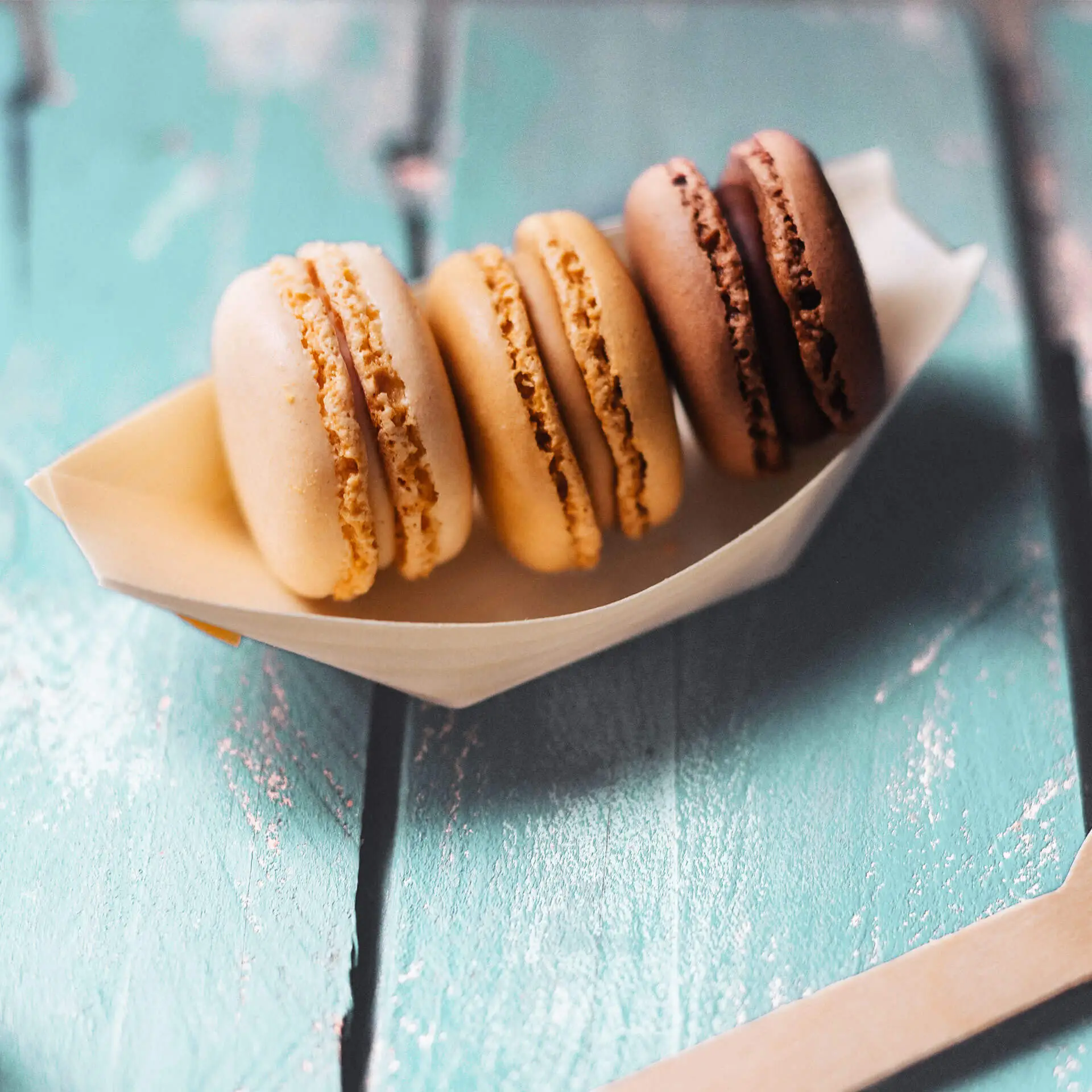 Quatre macarons dans un support en forme de bateau, de couleur beige, marron clair, marron foncé et chocolat sur une surface texturée.