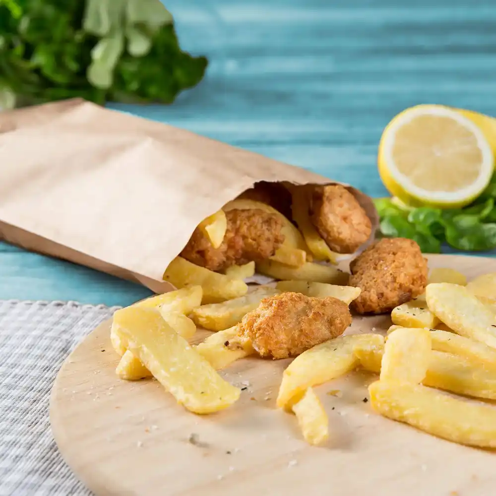 Sac en papier avec de la nourriture frite qui déborde, comprenant des frites et plusieurs morceaux dorés, avec du citron et des herbes en arrière-plan.