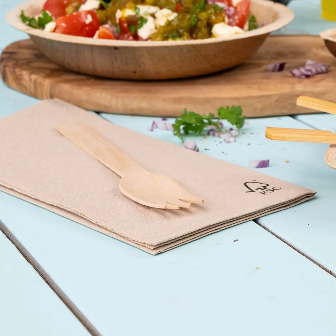 Fourchette en bois posée sur une serviette beige pliée, avec un bol de salade en arrière-plan sur une table bleu clair.