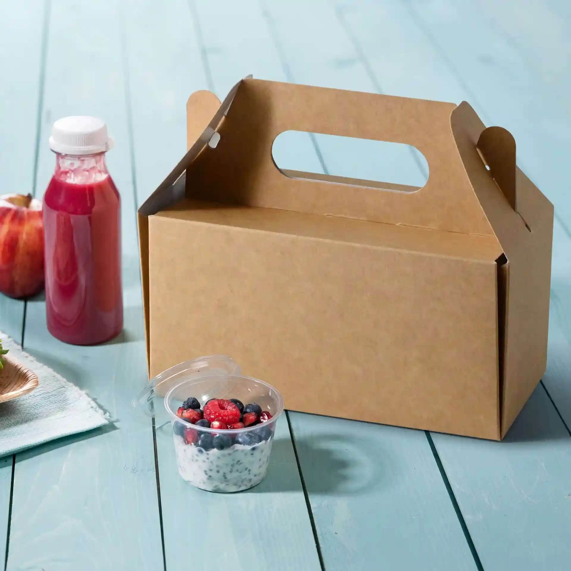 Boîte à pâtisserie en carton avec poignée, 26 x 17 x 9 cm, à côté d'une coupe de fruits et d'une bouteille de jus sur une surface en bois bleu.