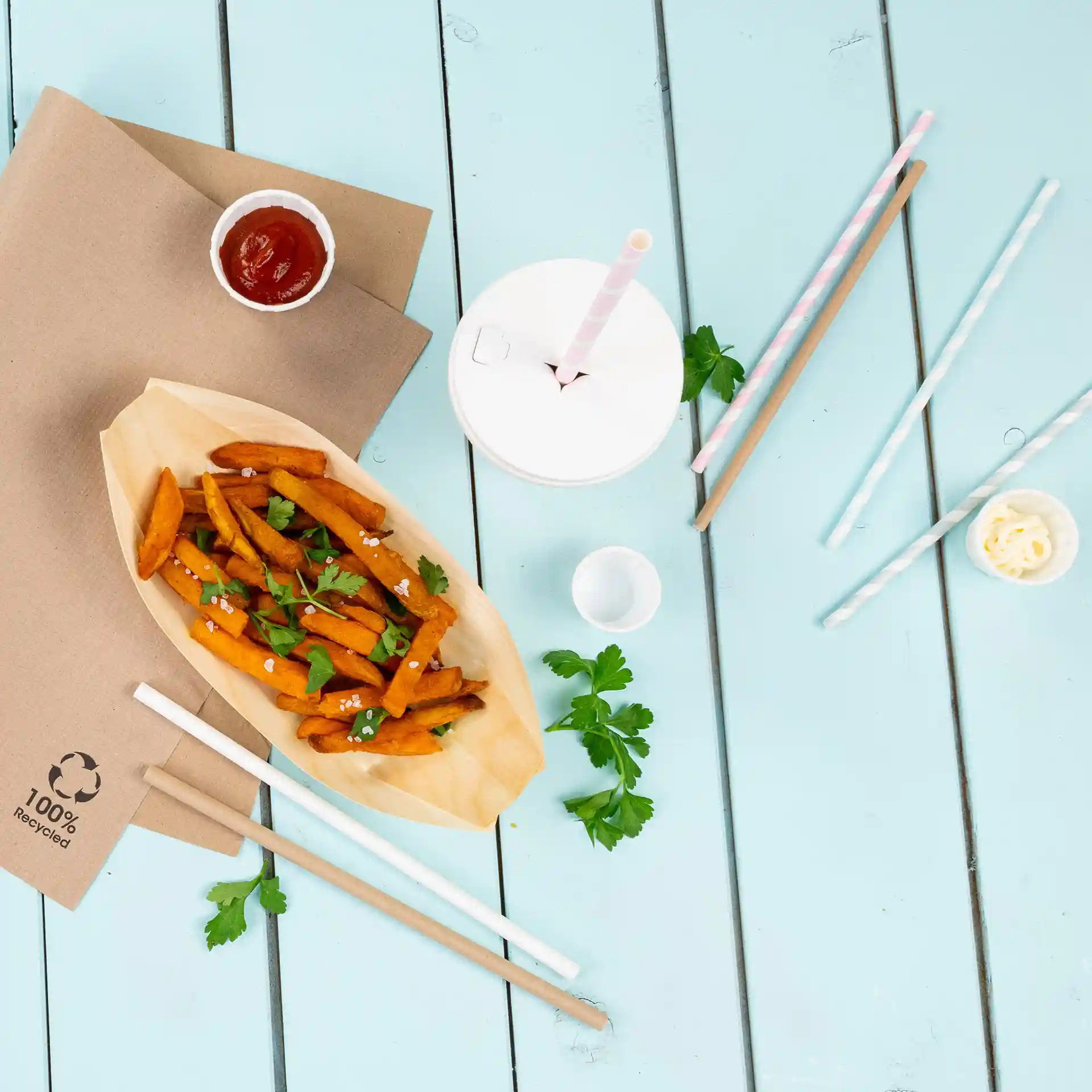 Image de frites de patate douce dans un bateau en bois avec du persil, du ketchup, un gobelet et des pailles en papier sur une table bleue.