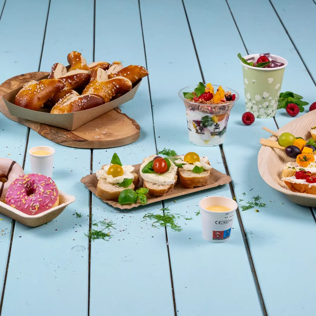 Assortiment de snacks comprenant des bretzels, des sandwiches garnis, un parfait au yaourt, des donuts et des brochettes de fruits sur une surface en bois bleu.