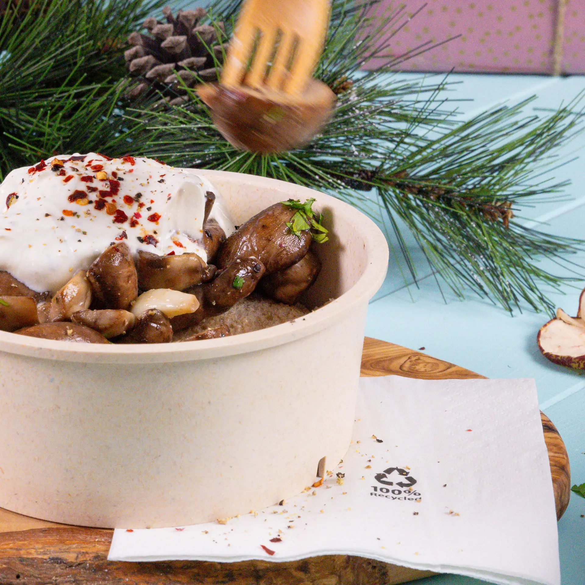 Un bol durable de champignons mélangés surmonté de yaourt et de flocons de piment, avec une fourchette en bois soulevant un morceau ; serviette en dessous.