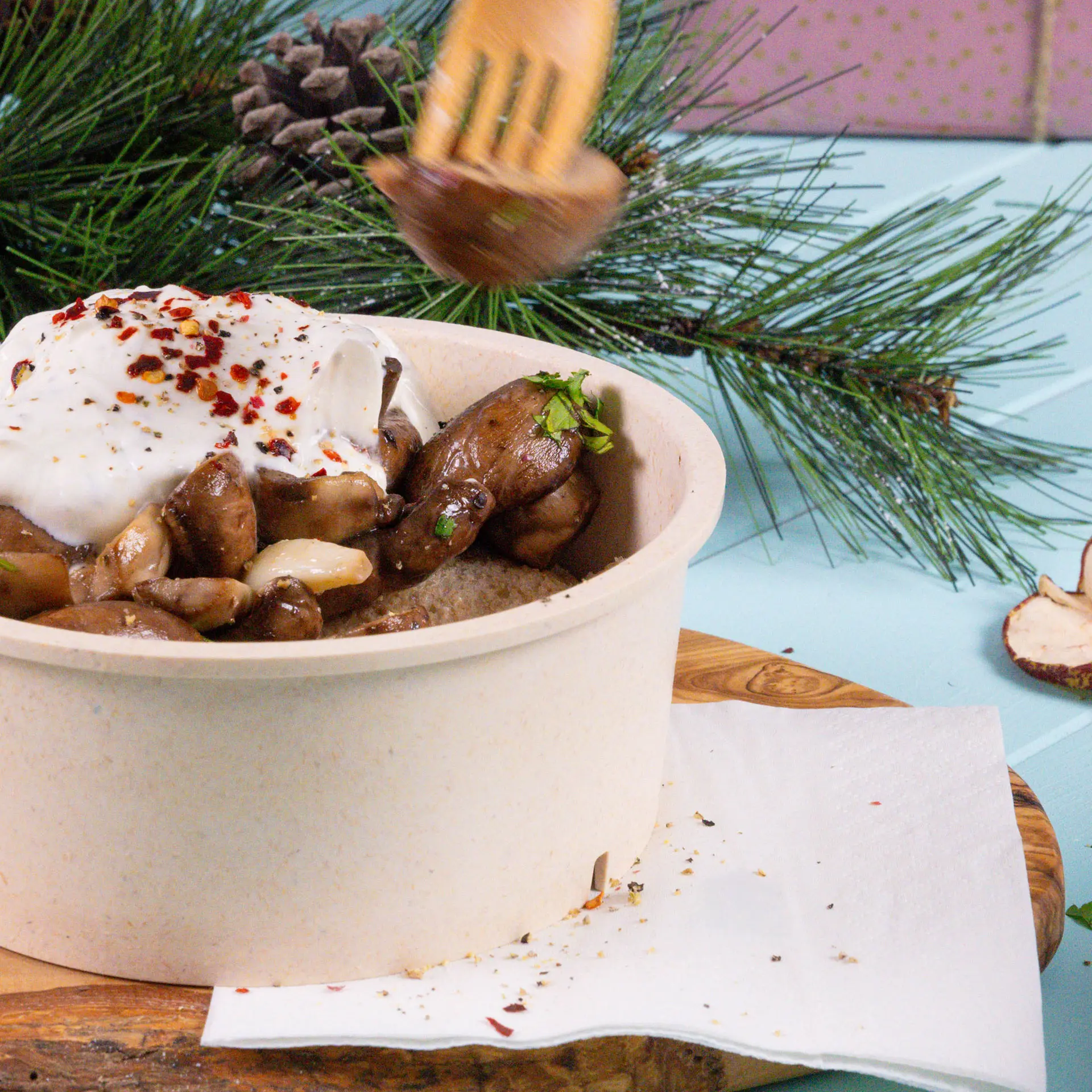 Bol avec des champignons et du yaourt, garni de flocons de piment rouge, placé sur une planche en bois avec une serviette blanche à côté.