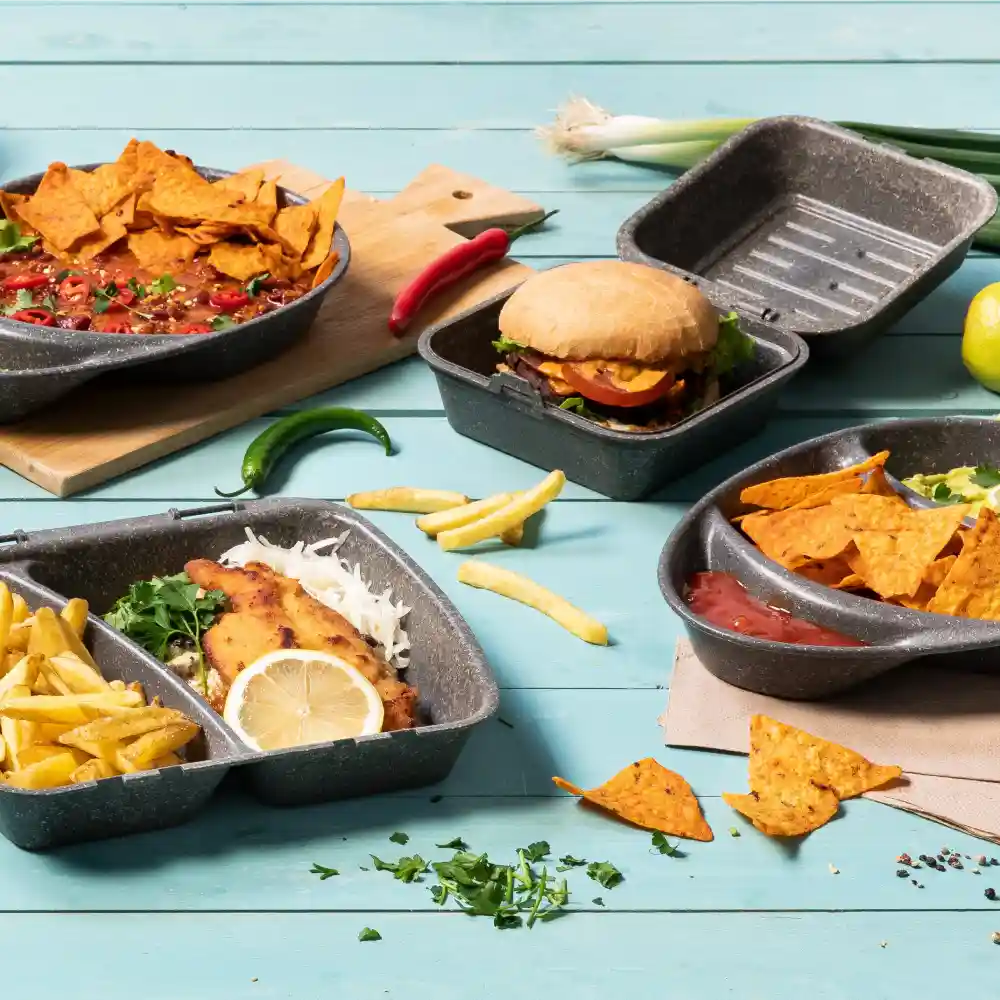 Variété d'aliments dans des contenants réutilisables, y compris des nachos, un burger, des frites et du poisson avec du citron, sur une surface bleue.