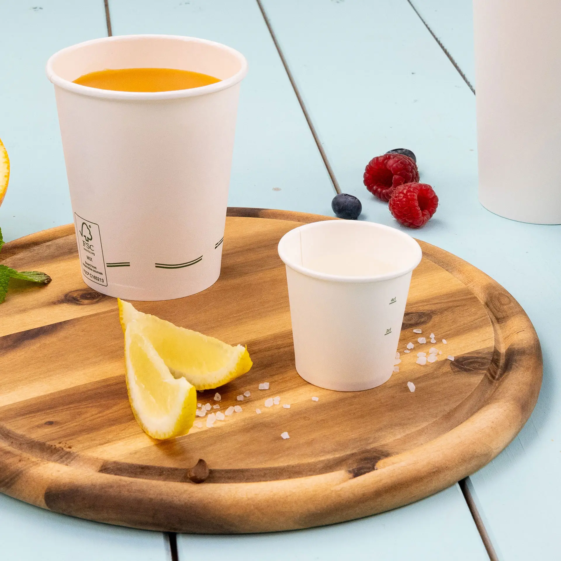 Image d'un plateau en bois avec un gobelet en papier blanc rempli de liquide orange, un plus petit gobelet en papier, des quartiers de citron et du sel.